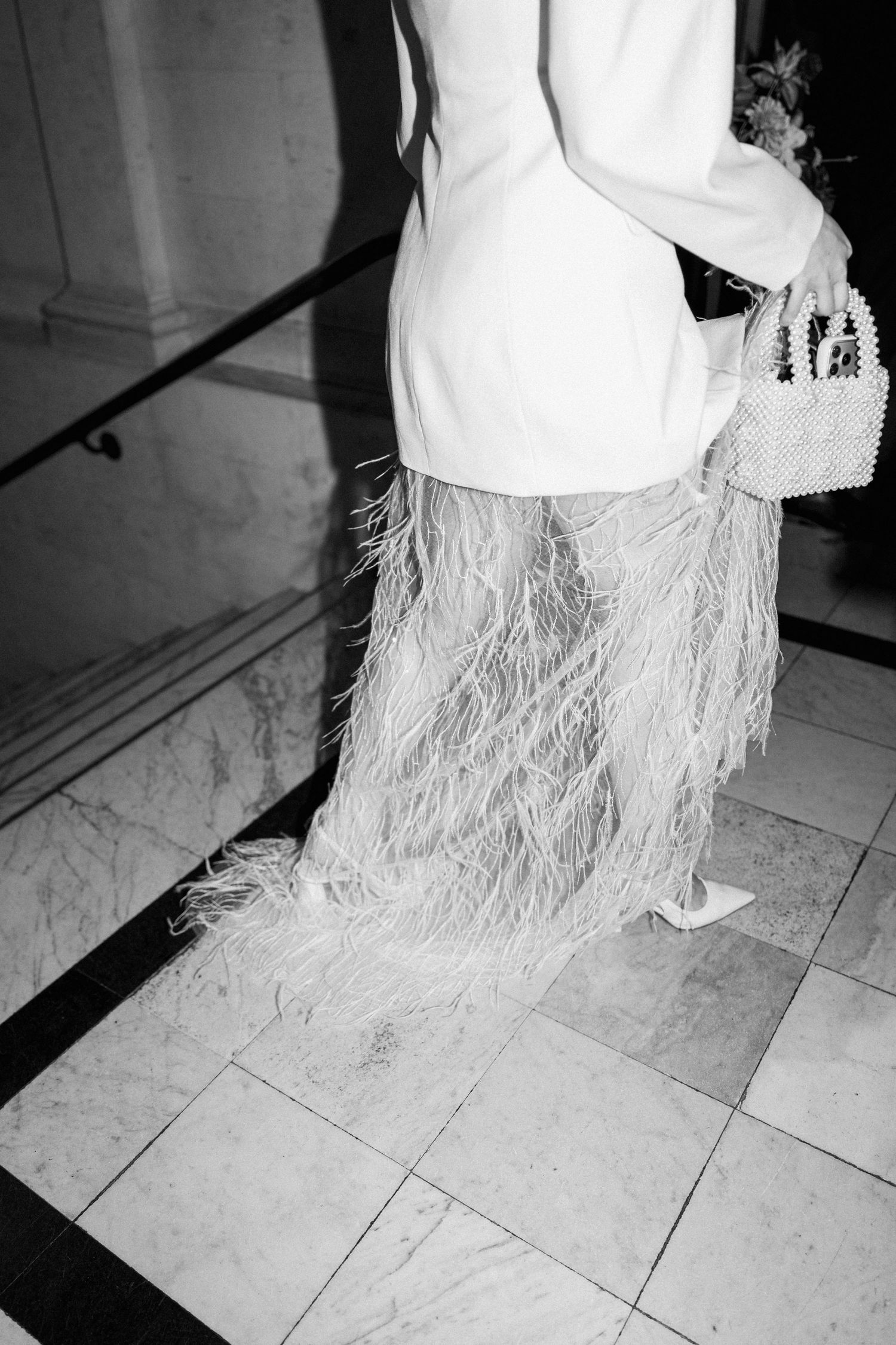 Black and white photo of bride in white feathered dress carrying pearl handbag on marble stairs.
