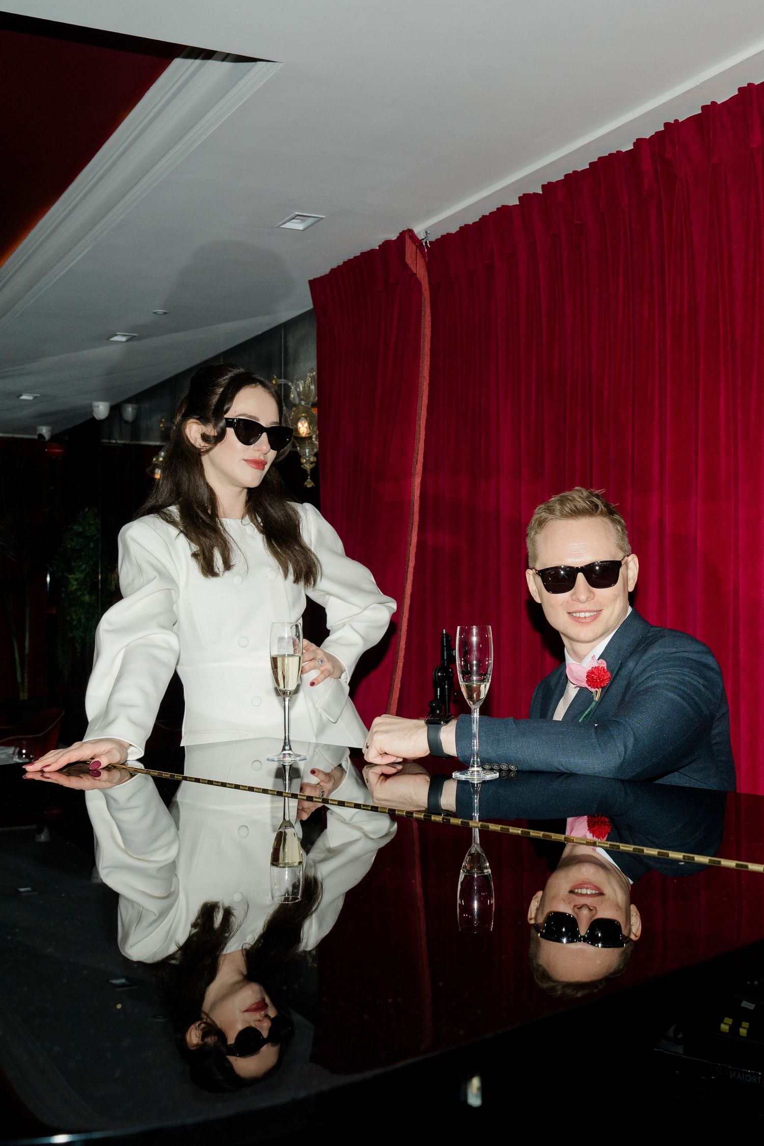 Stylish bride and groom in sunglasses toast champagne at mirrored table with red velvet backdrop.