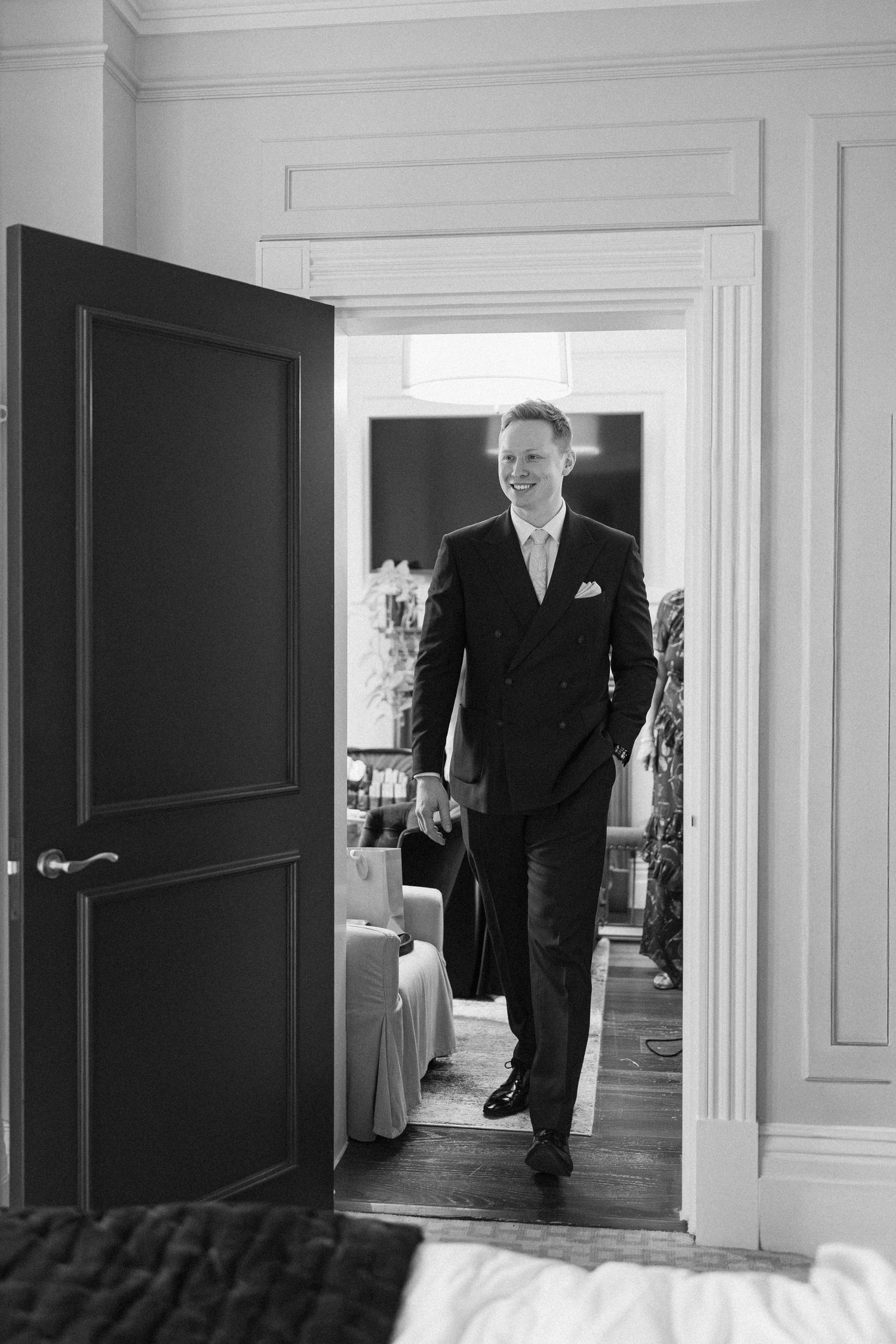 Smiling groom in black double-breasted suit walking through doorway in black and white photo.