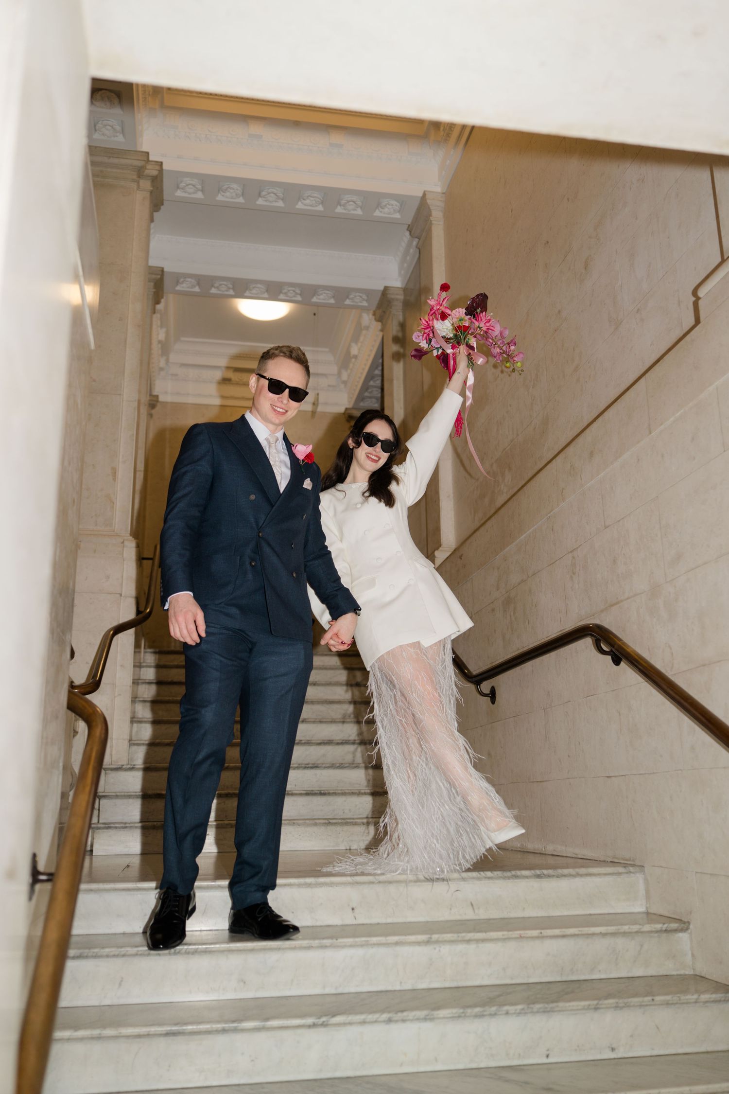 Joyful newlyweds in sunglasses descending grand marble staircase, bride raising colorful bouquet.