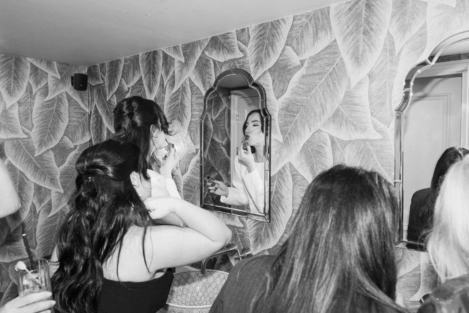 Black and white photo of bride checking hair in arched mirror with leaf-patterned wallpaper backdrop.