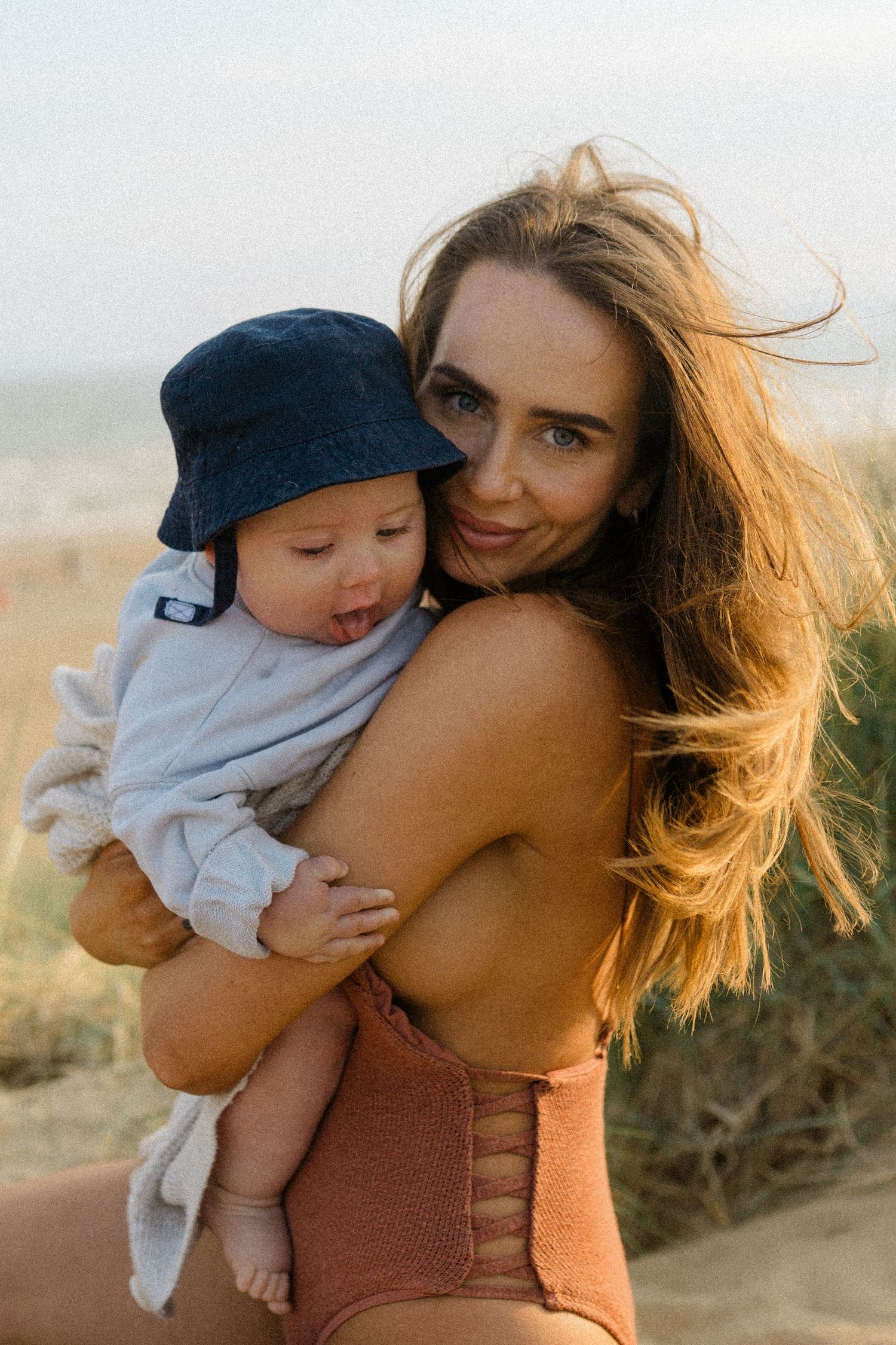 Young mother in orange swimsuit holds baby wearing blue bucket hat on a sunny beach.