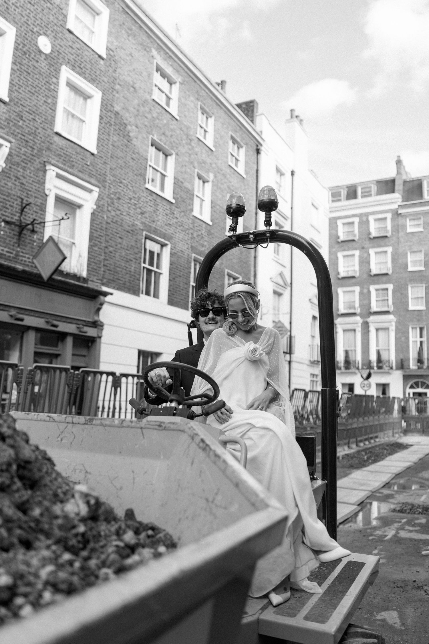 A couple in wedding attire pose playfully on a yellow construction vehicle in an urban setting.