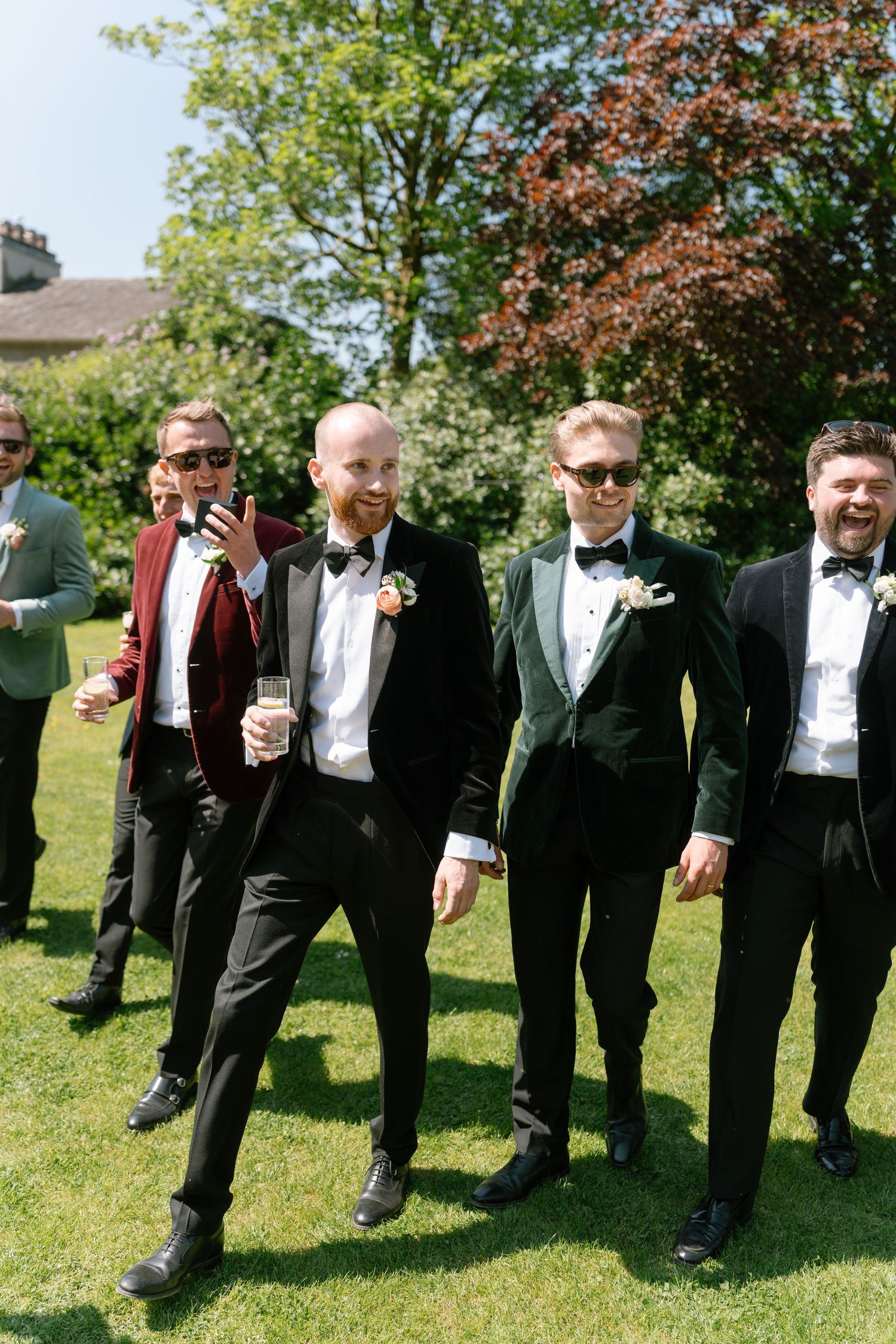 Group of formally dressed men stand in a row on sunny lawn holding drinks at wedding event.