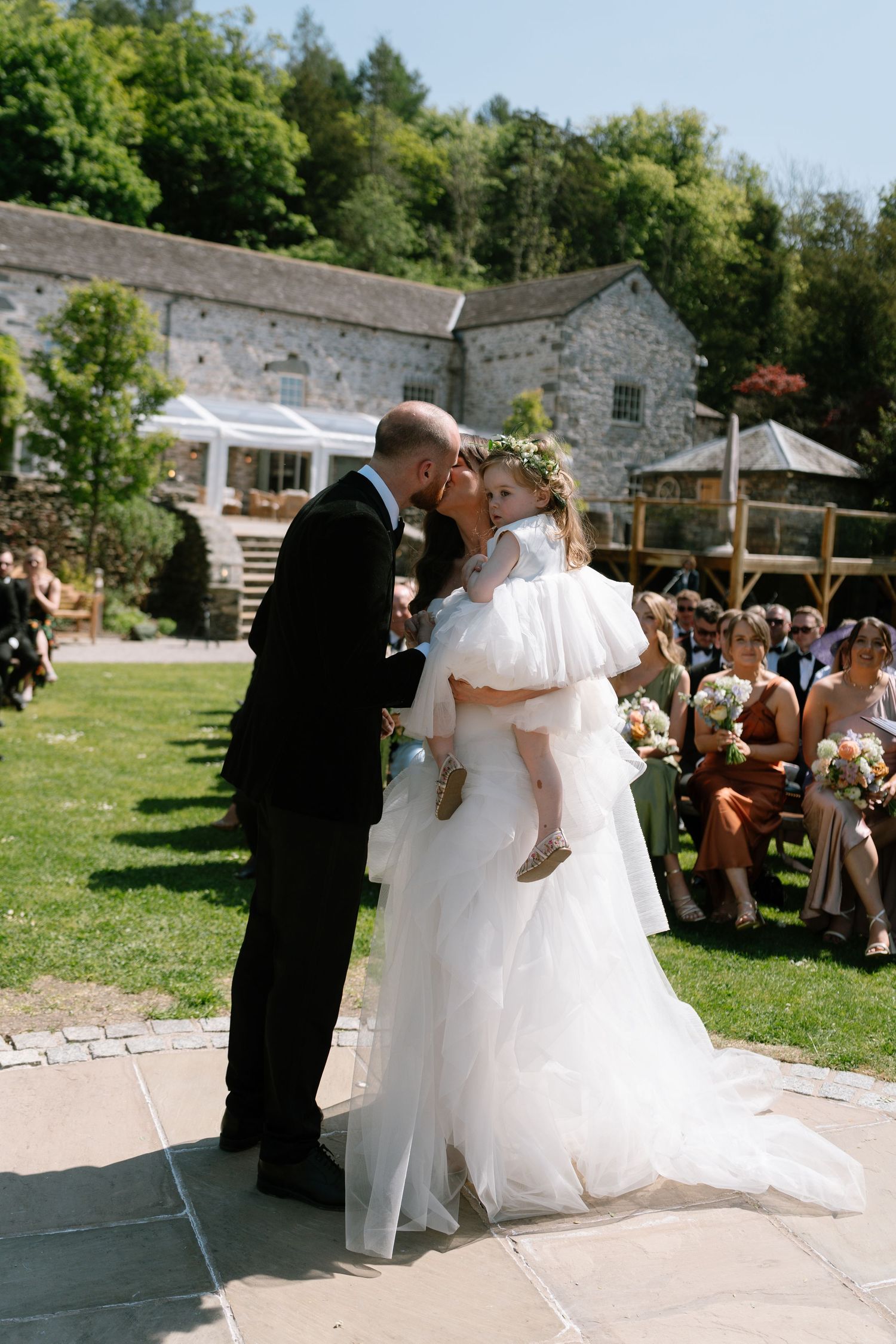 Sunny outdoor wedding celebration with stone building backdrop and gathered guests.