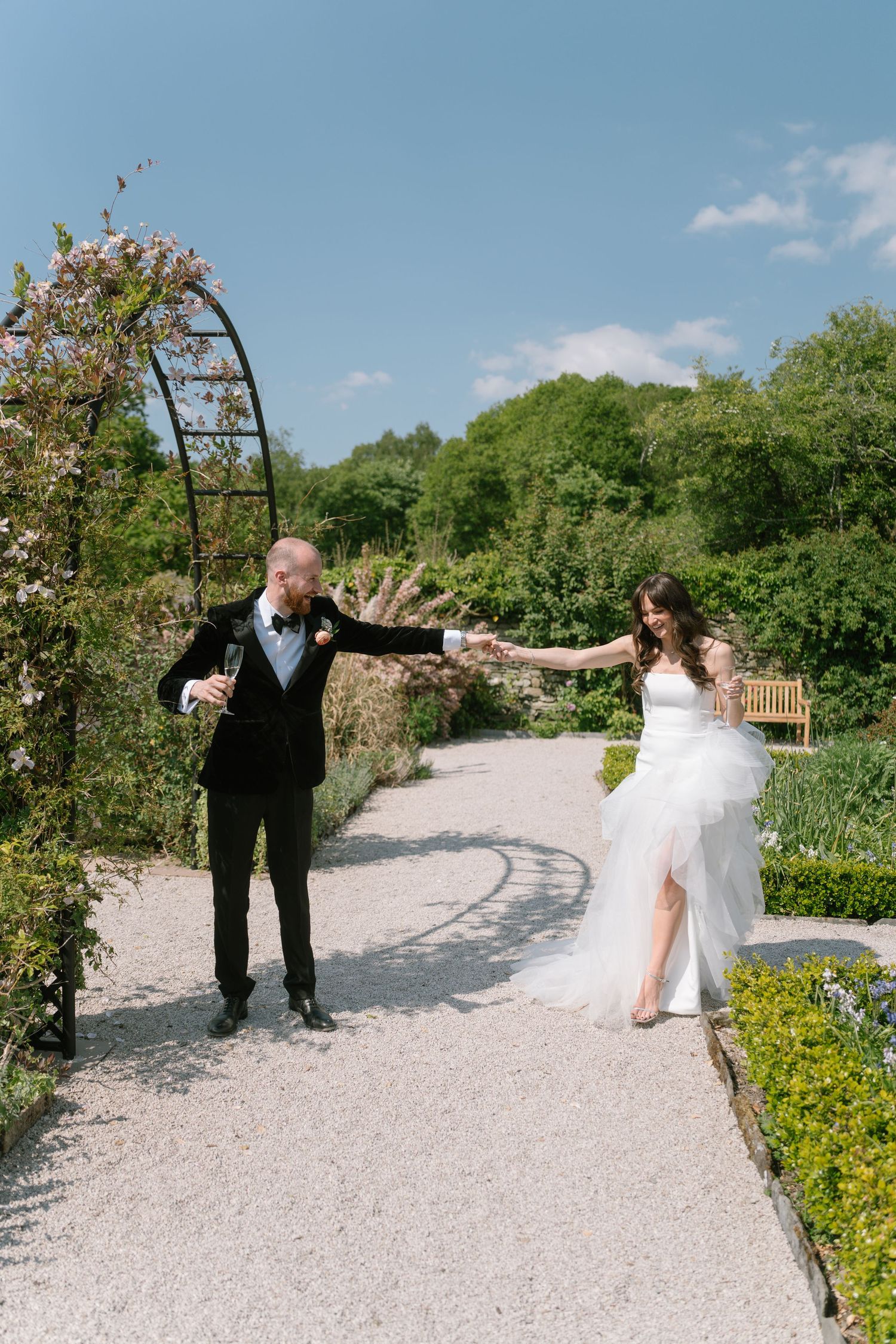 A playful wedding photoshoot on a winding garden path with yellow wildflowers and greenery.