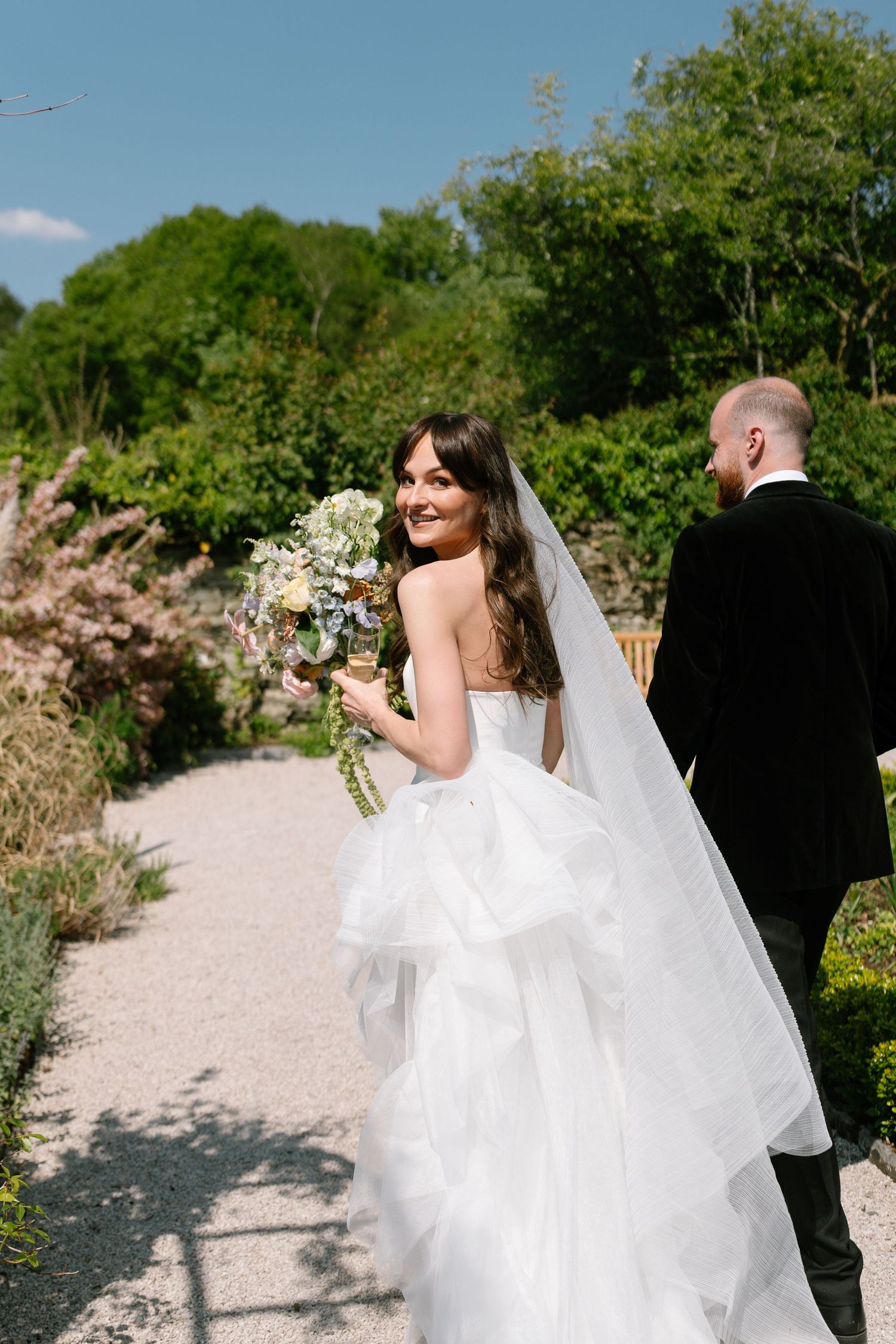 Wedding party walking through a sunlit garden path with flowing white dress.