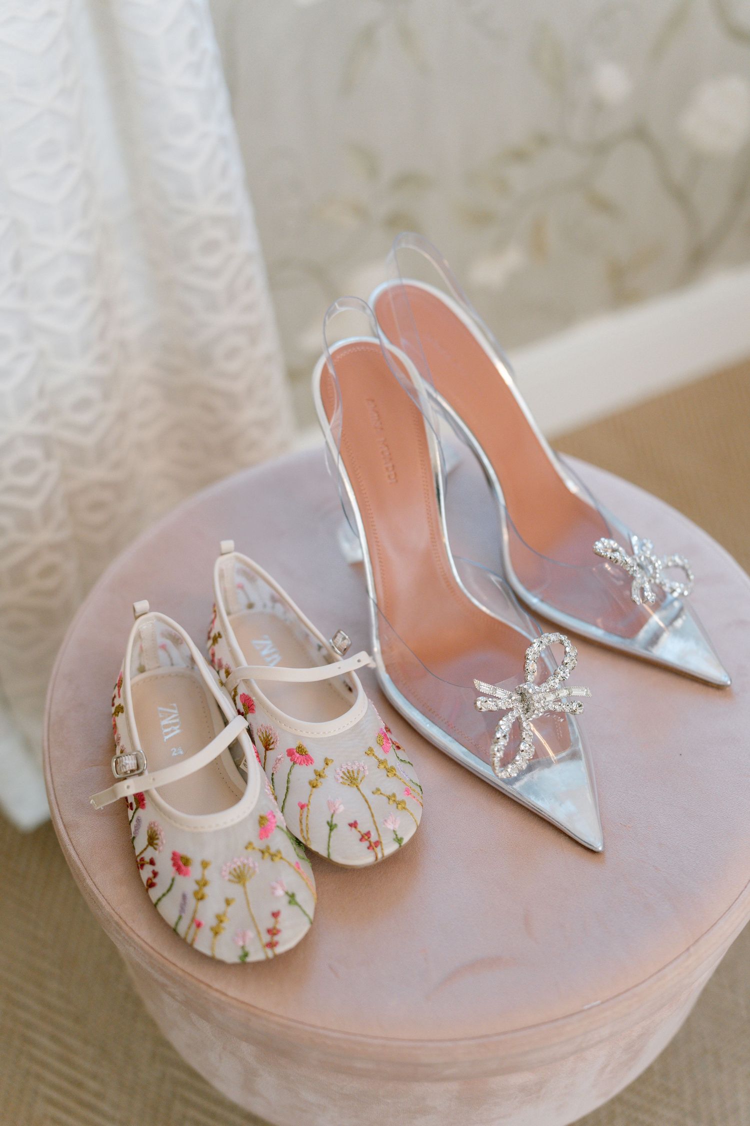 Floral patterned baby shoes next to silver heels on a pink cushioned surface.