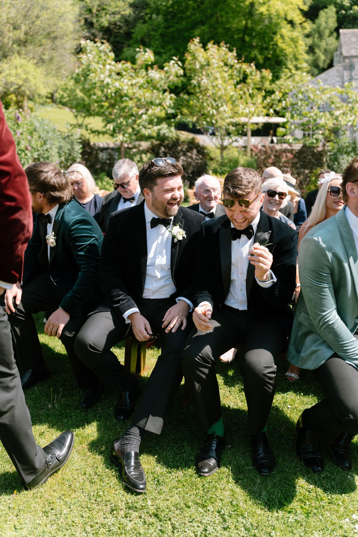 Row of formally dressed guests sitting on benches in a sunny garden setting.