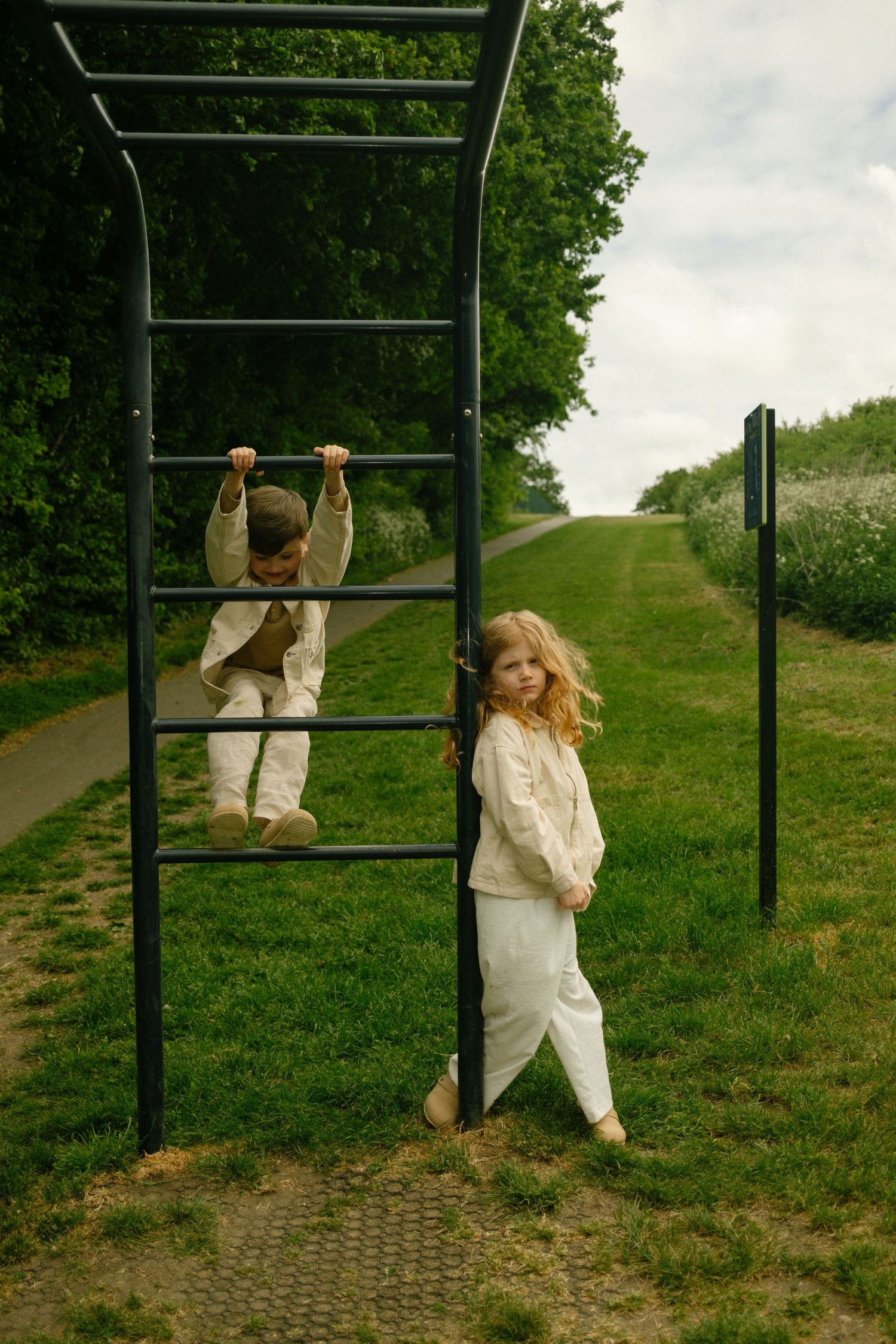 Children in white clothing play on outdoor exercise equipment along a green path.