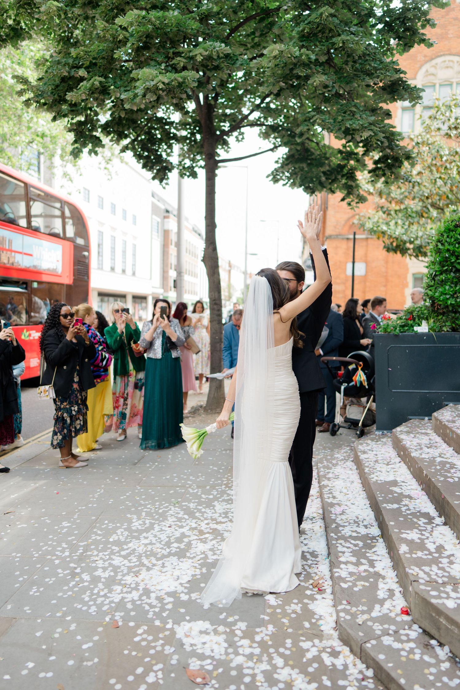 Bride and groom kiss outside London church surrounded by confetti, guests, and a red double-decker bus.