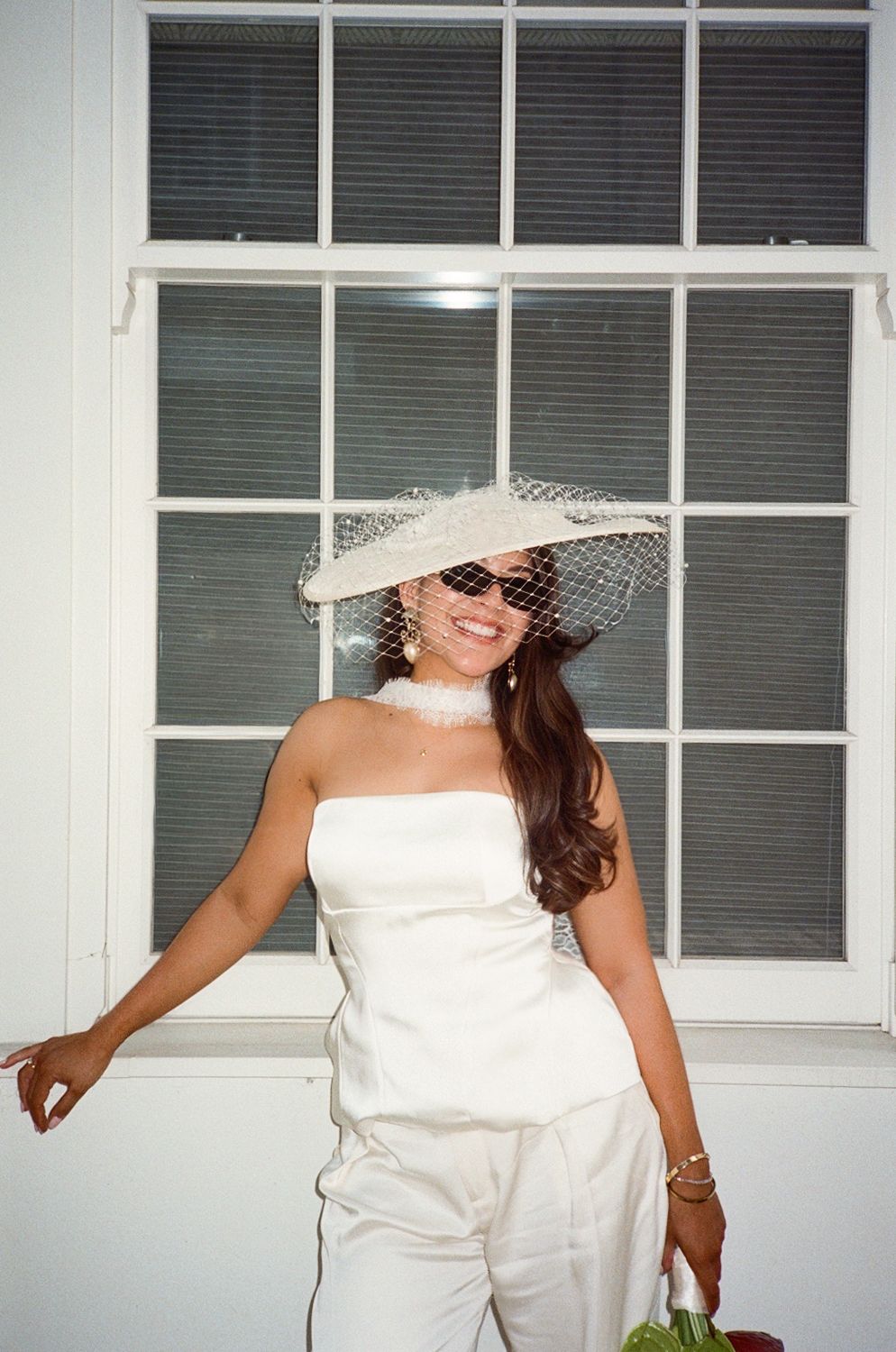 Woman in white strapless jumpsuit and wide-brim hat with veil netting smiles in front of window.