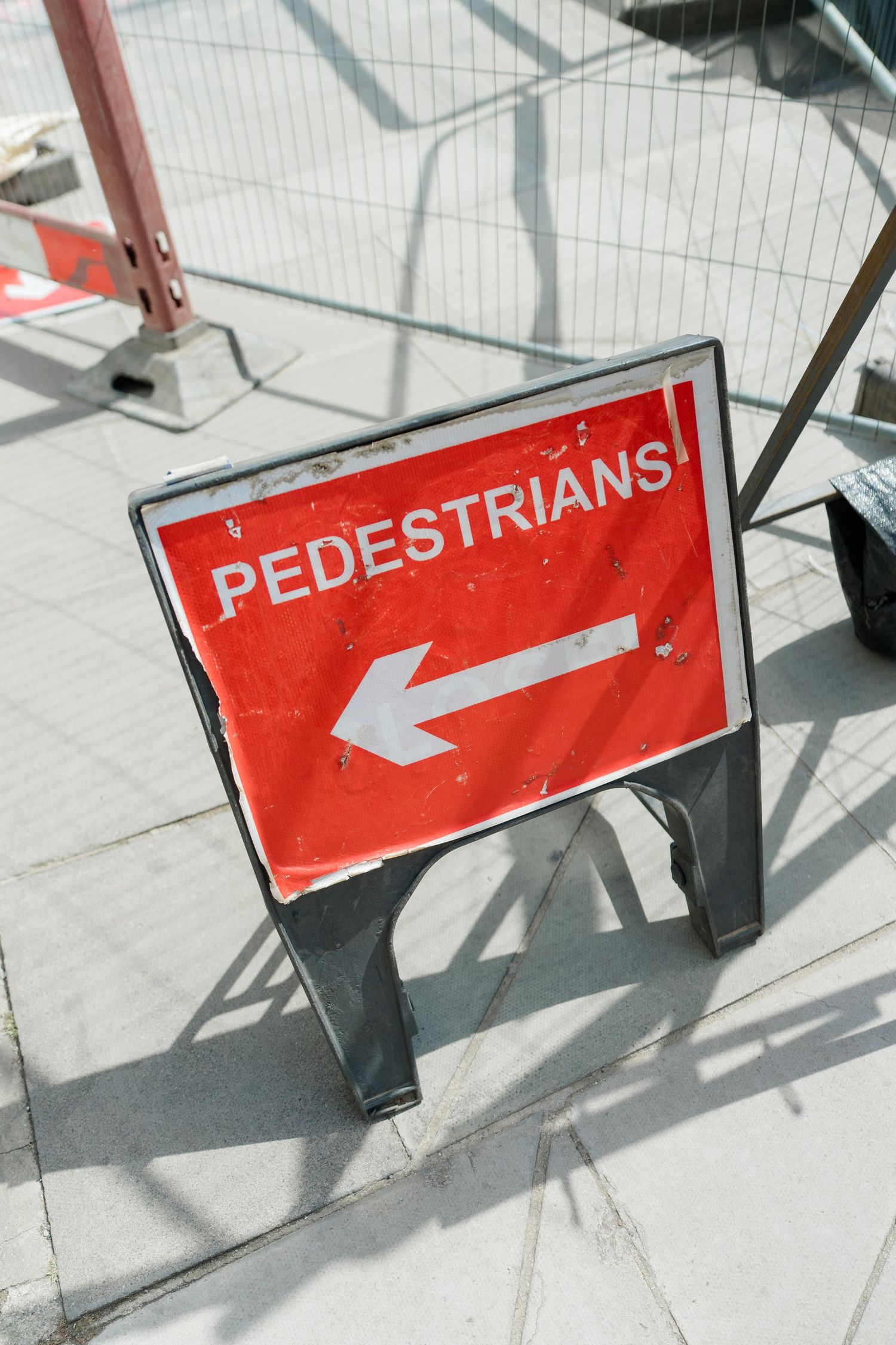 Red pedestrian directional sign with white arrow on city sidewalk near construction barriers.