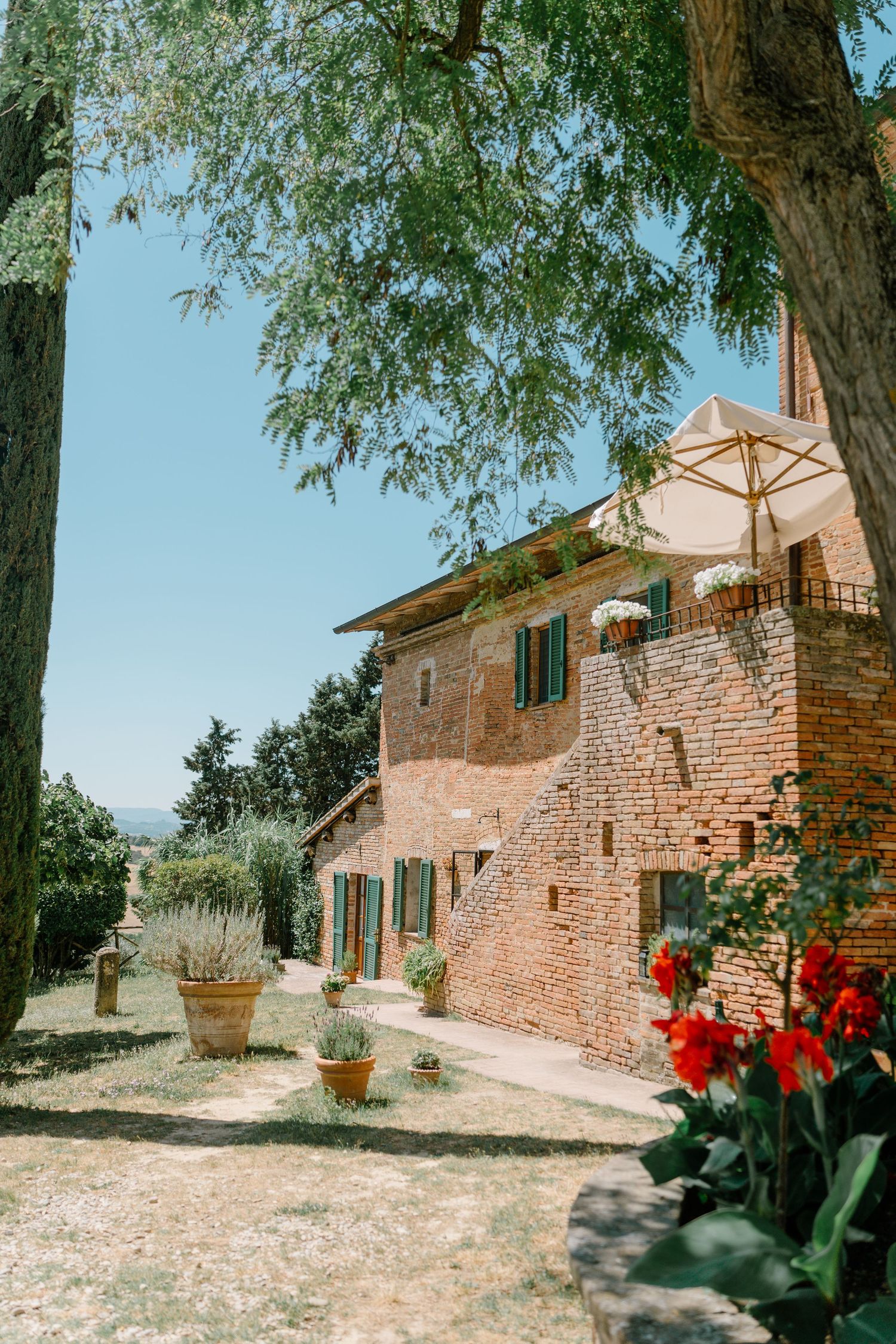 A rustic stone farmhouse in Tuscany with red tulips blooming in the garden and trees providing natural shade.