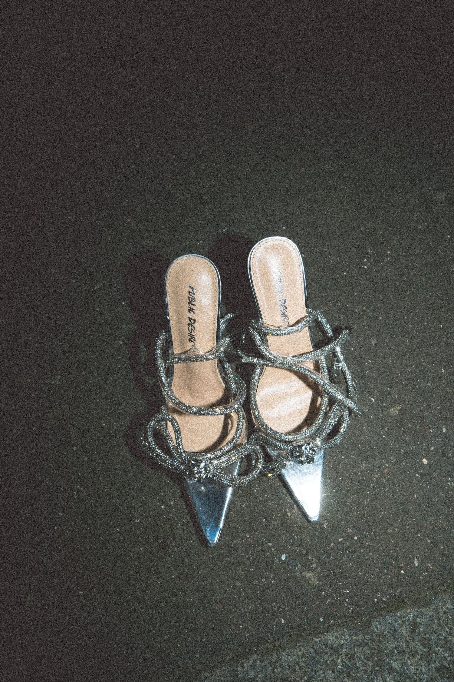 A pair of silver metallic pointed-toe heels with straps lies on a dark textured surface.