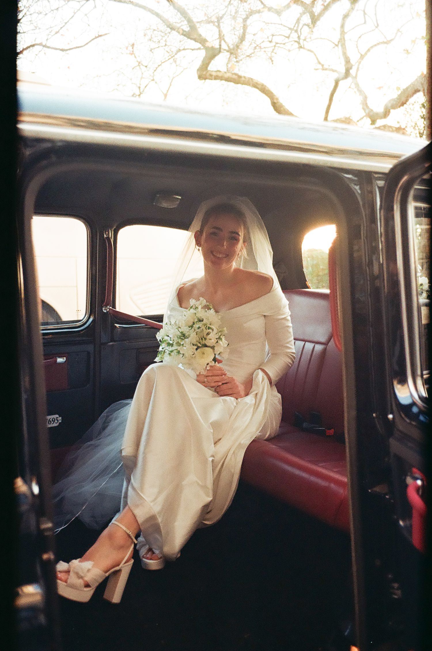 A person in white attire sits inside a vintage car interior with dark upholstery during what appears to be a wedding.