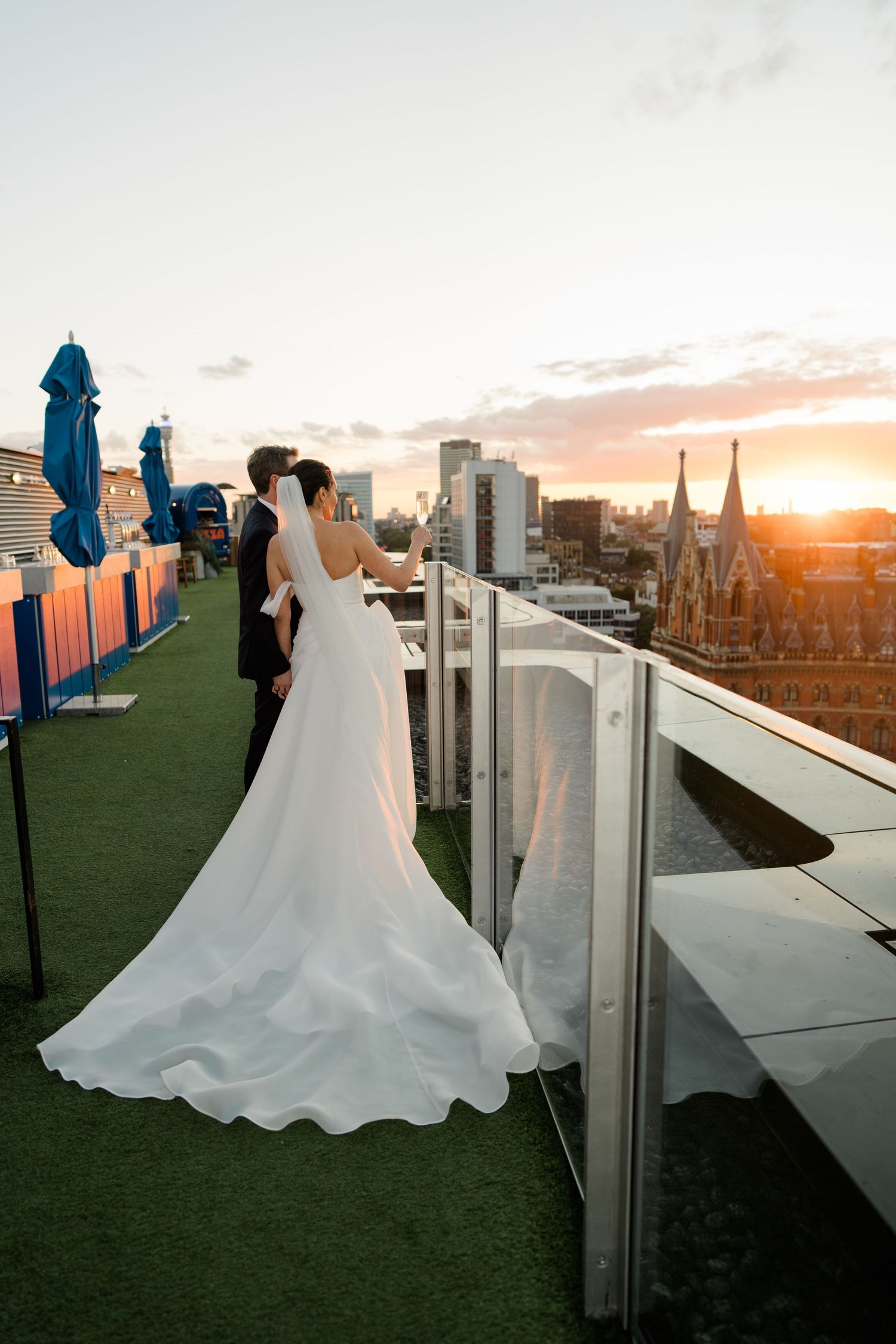 A bride in a flowing white gown poses against a sunset cityscape on a rooftop terrace.