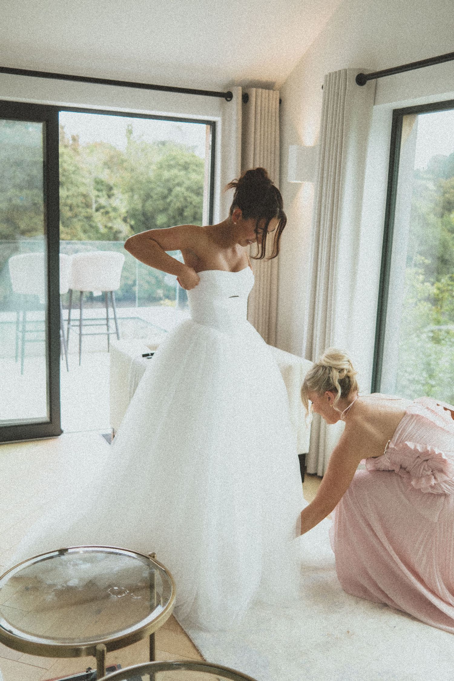 A bride in a white wedding gown twirls and dances in a bright sunlit room with large windows.