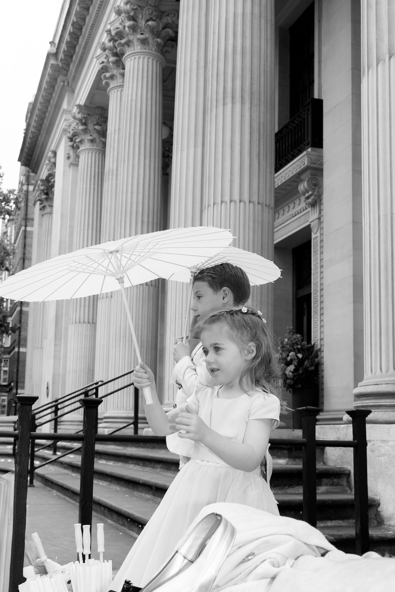 Young flower girls in white dresses hold parasols while sitting on steps with classical columns.