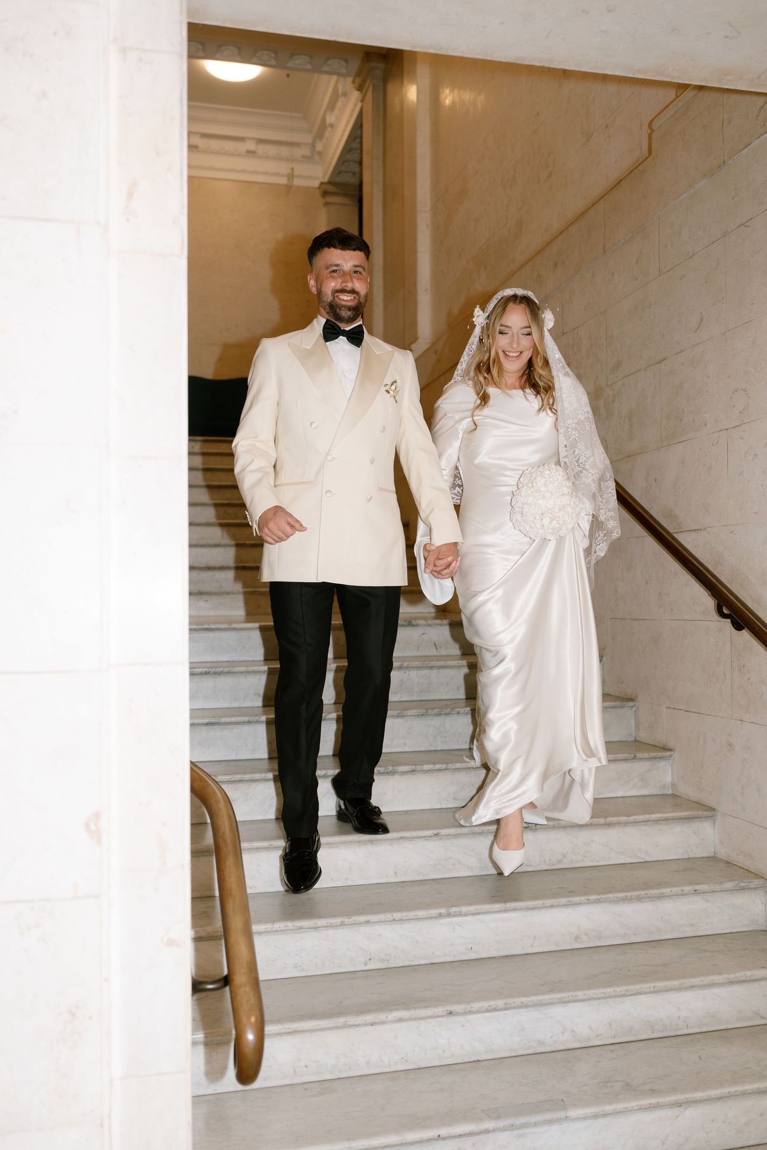 A couple descends elegant marble stairs during their wedding, with the bride wearing a flowing white dress.