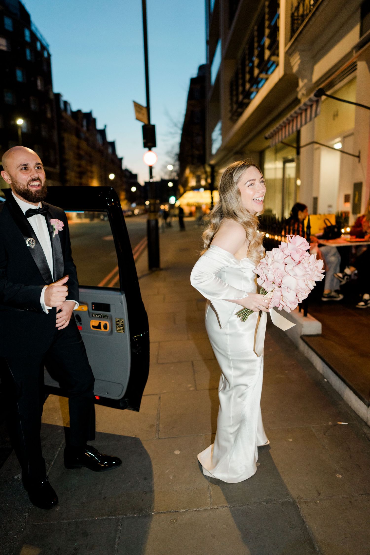 Wedding couple on city street at dusk with pink bouquet and formal attire.