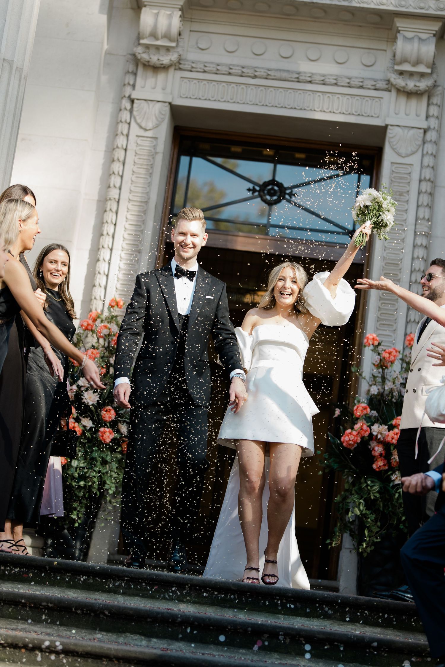 Wedding celebration outside historic building with confetti flying through the air.