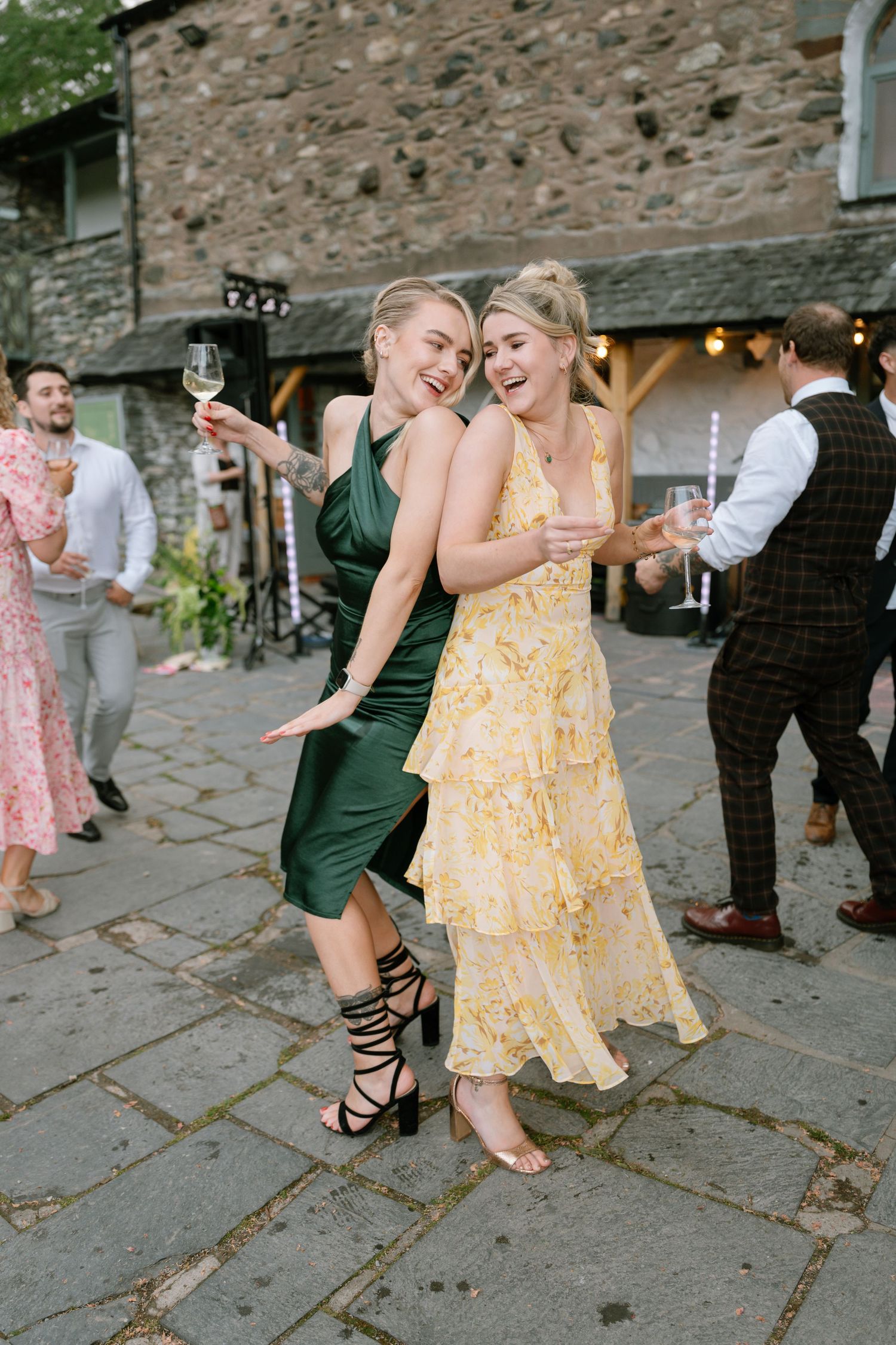 Wedding guests in colorful dresses enjoying drinks and dancing at an outdoor stone venue reception.