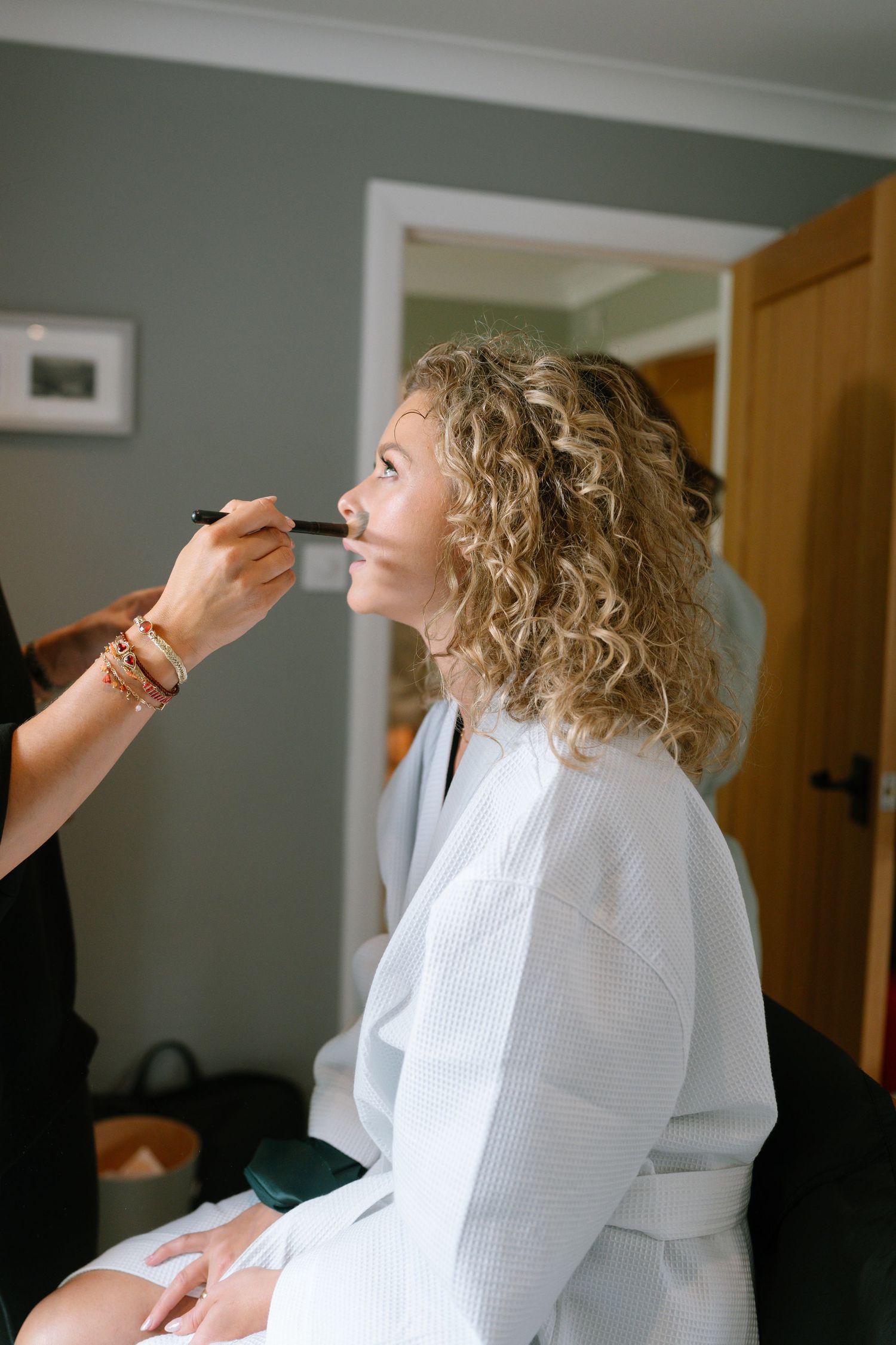 Person in white robe getting makeup applied while sitting in preparation room.