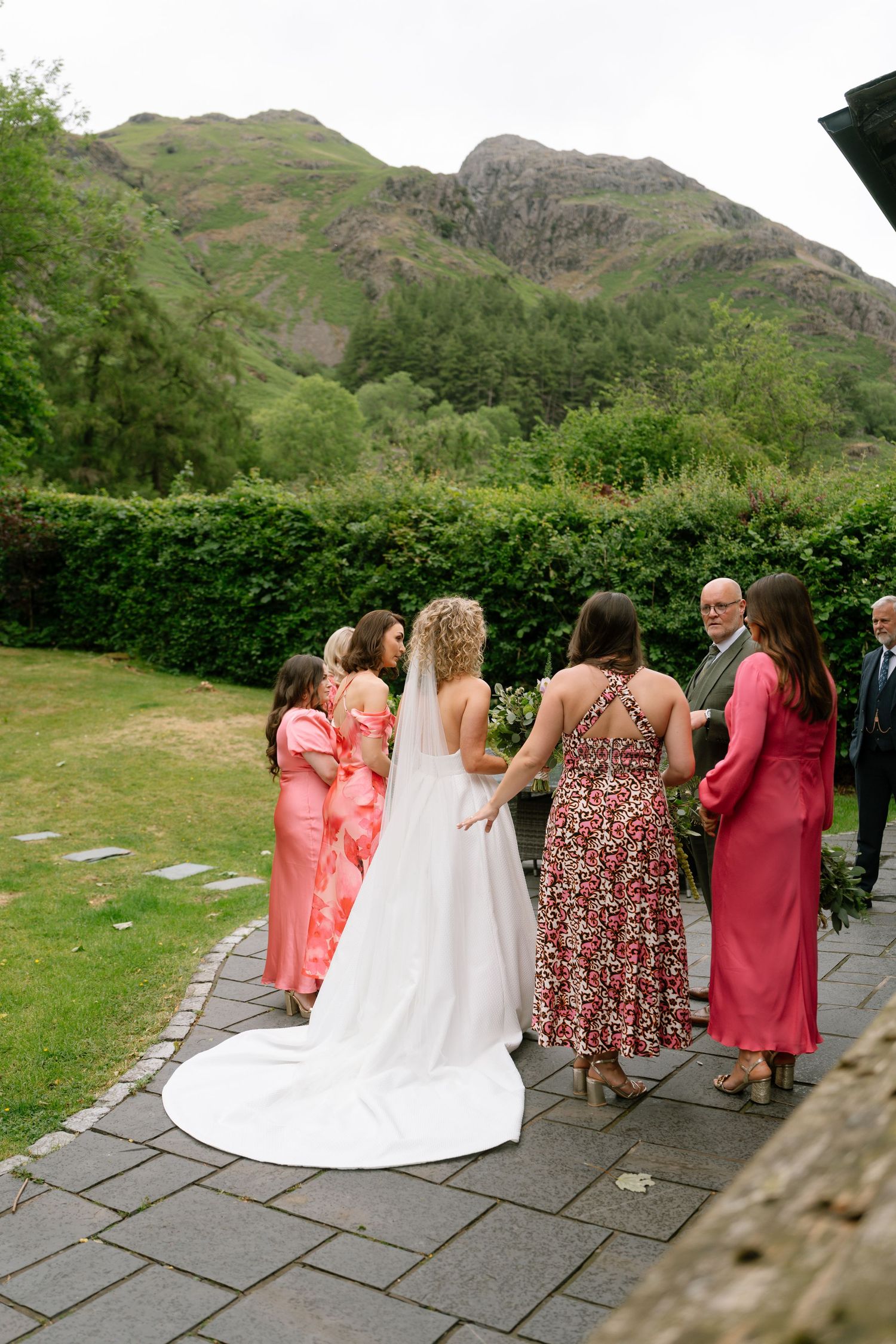 Wedding party in coral dresses and white gown pose against scenic mountain backdrop on stone pathway.