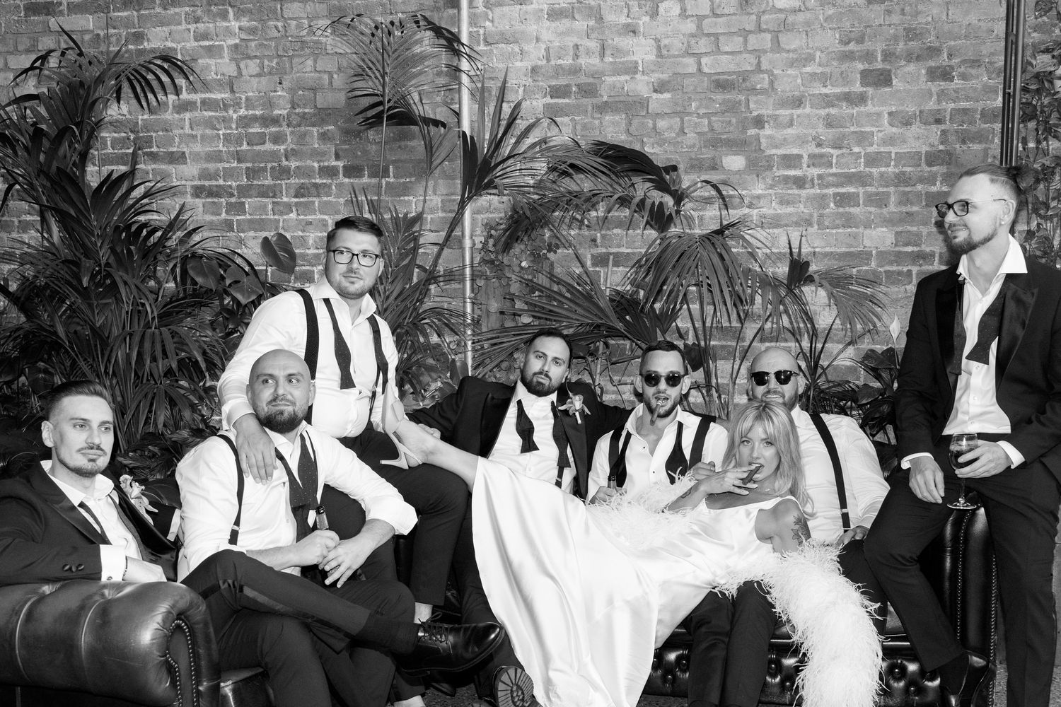 Wedding party lounges on leather couches against a brick wall backdrop in black and white.
