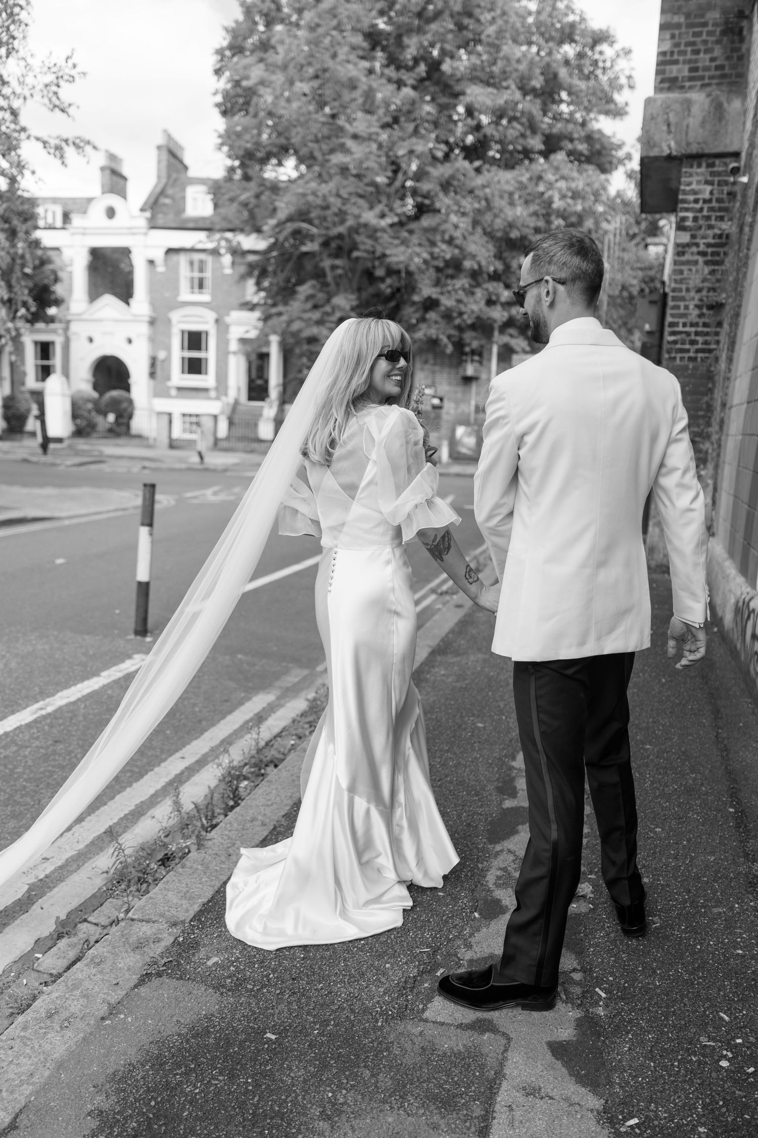 A bride in a flowing dress and groom walk along a street in black and white photography.