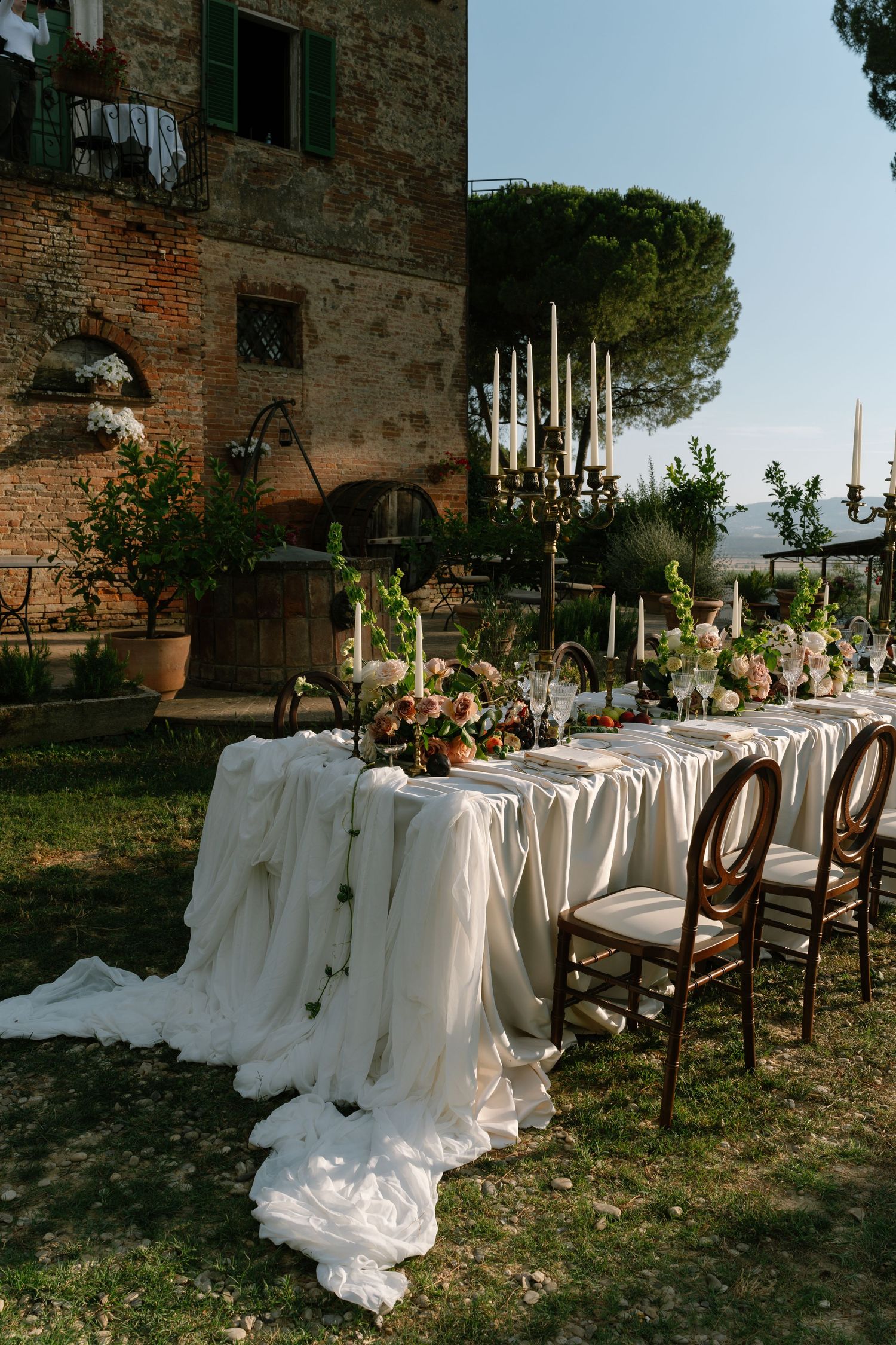 Elegant outdoor wedding reception table with white linens and rustic wooden chairs set against an old stone building.
