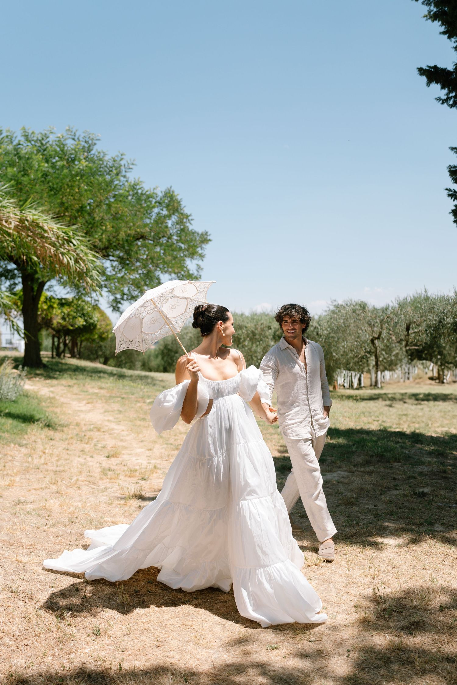 A couple shares a joyful moment walking through an olive grove with a white parasol.