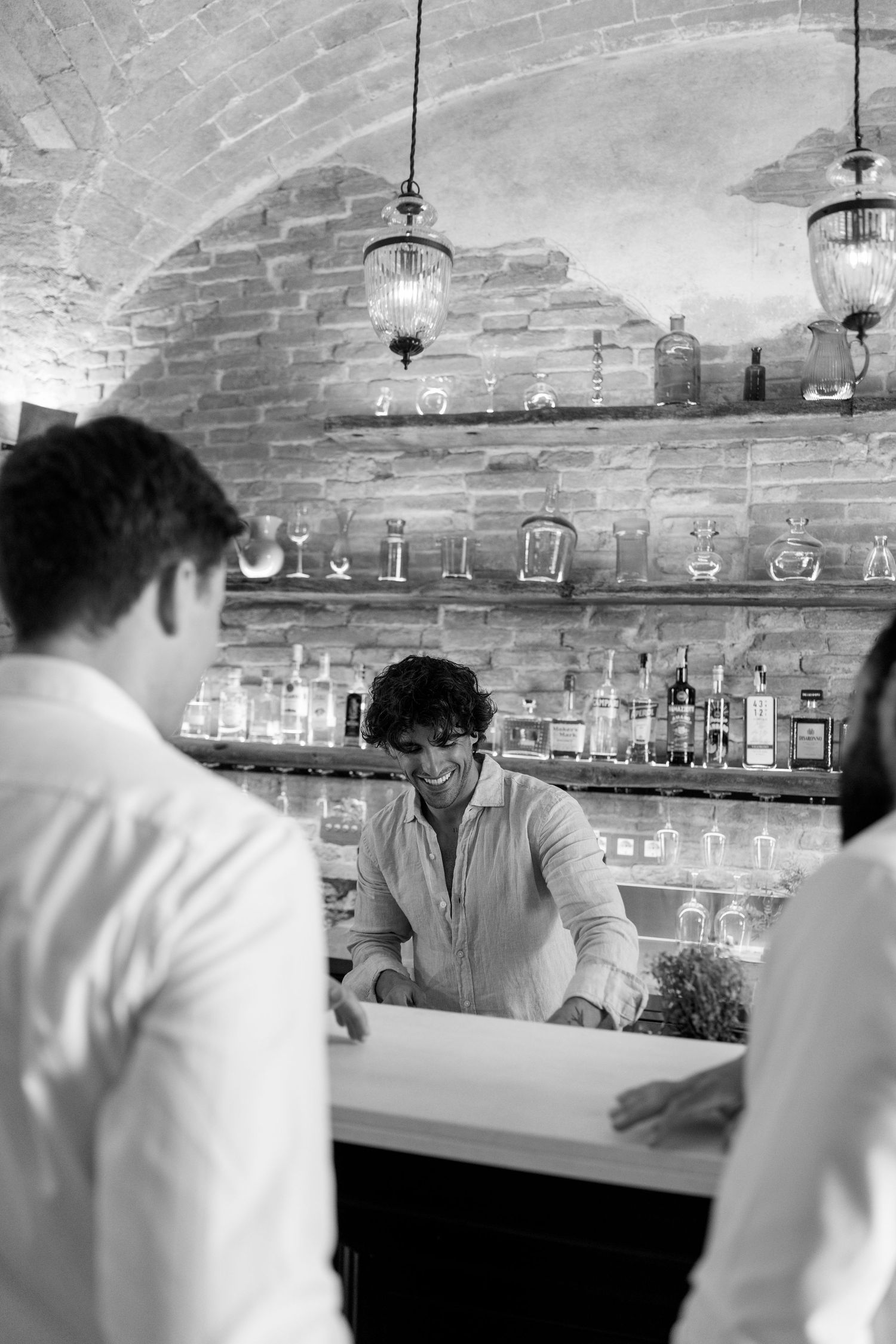 Bartenders mix drinks behind a rustic brick-walled bar with bottles lined up on display.