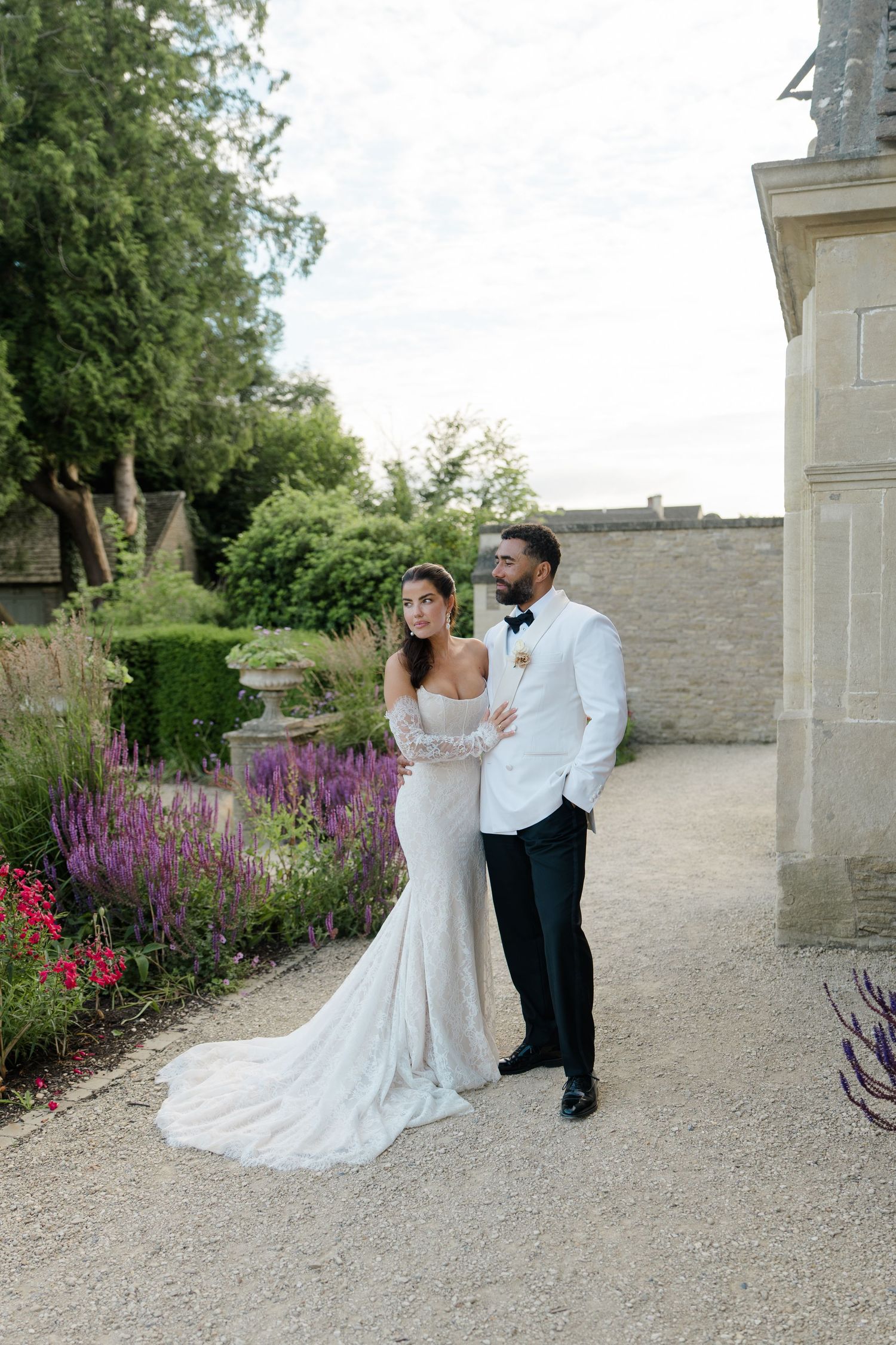 Bride in elegant off-shoulder lace gown and groom in white tuxedo posing beside stone building with garden flowers.