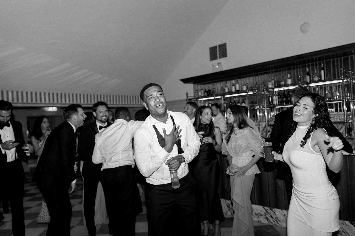Black and white photo of wedding guests dancing and celebrating energetically at indoor reception.