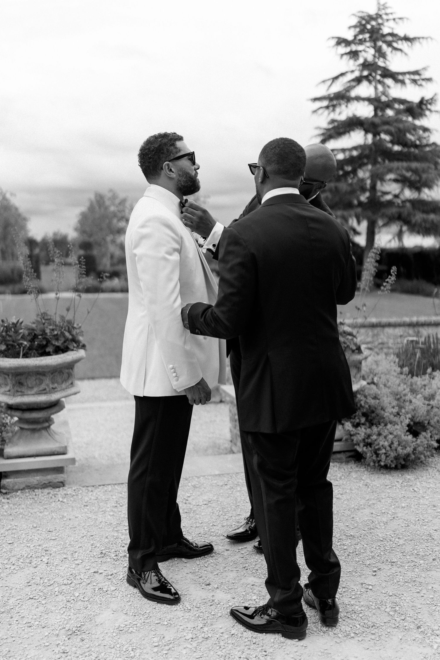 Black and white photo of groom in white jacket having bow tie adjusted by groomsmen outdoors.