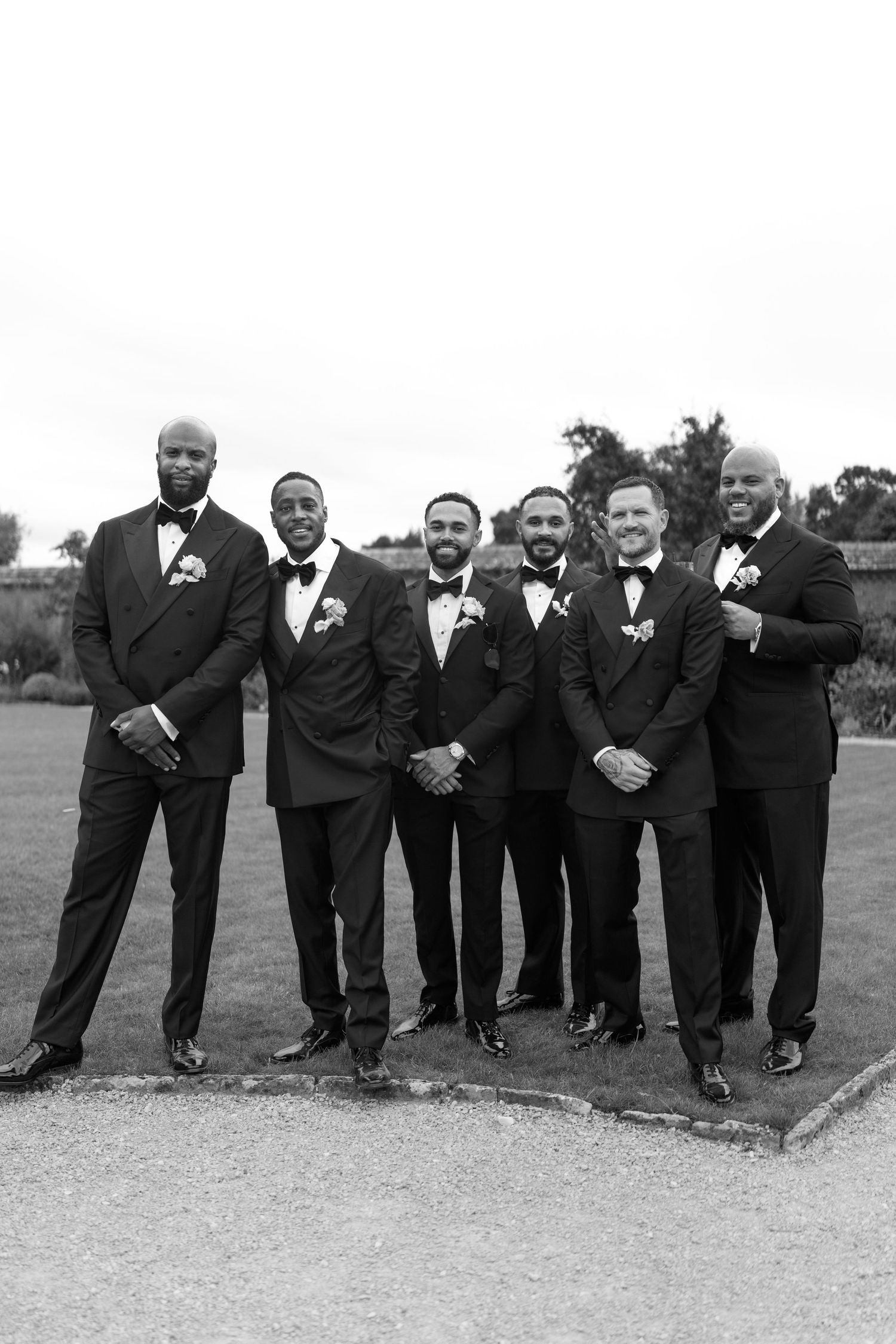 Groom poses with five groomsmen in black tuxedos outdoors in black and white.