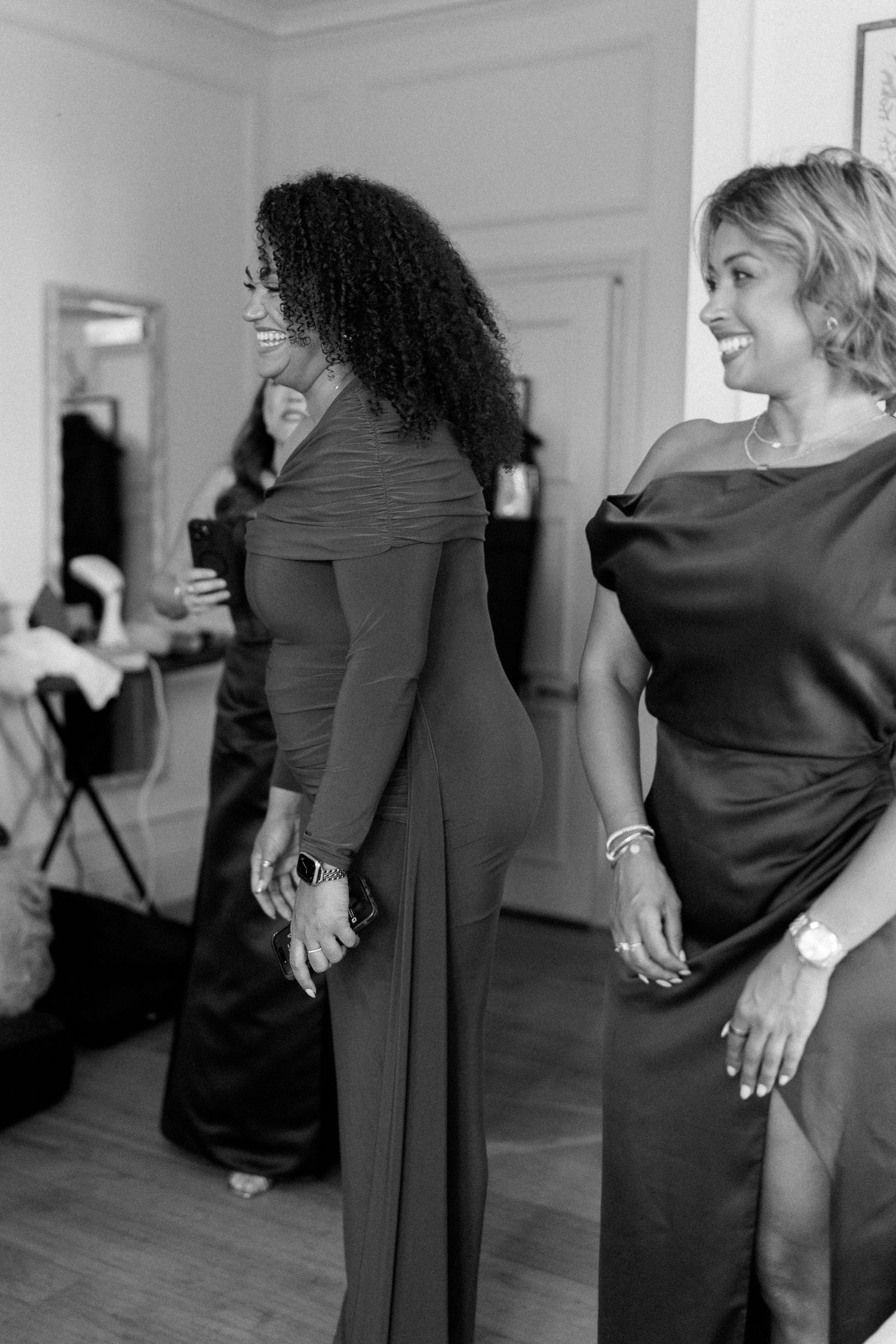 Two women in elegant dark gowns laugh together during wedding preparations in black and white photo.