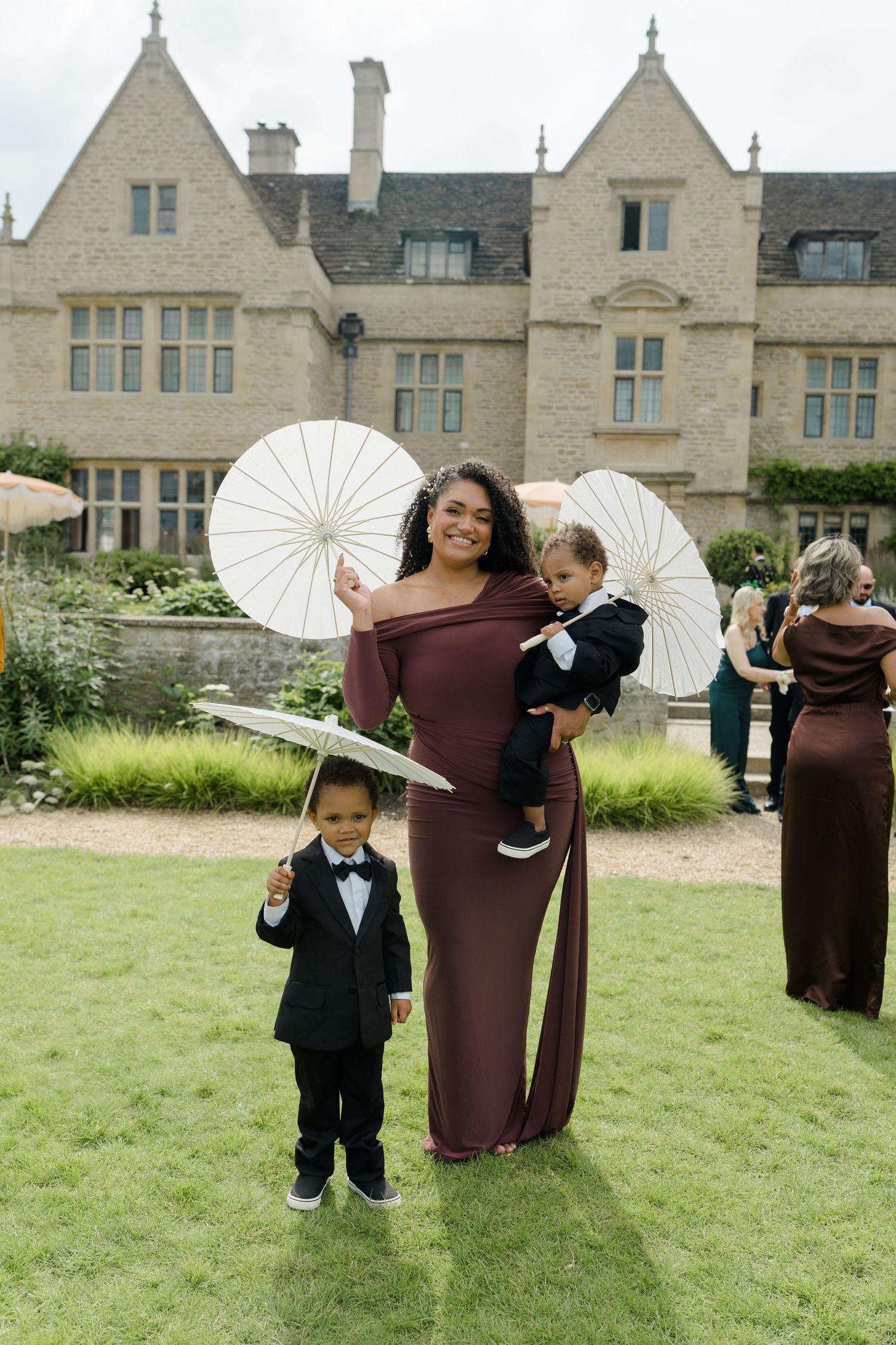 A woman in a burgundy gown holds two young boys in tuxedos with white parasols at a wedding venue.