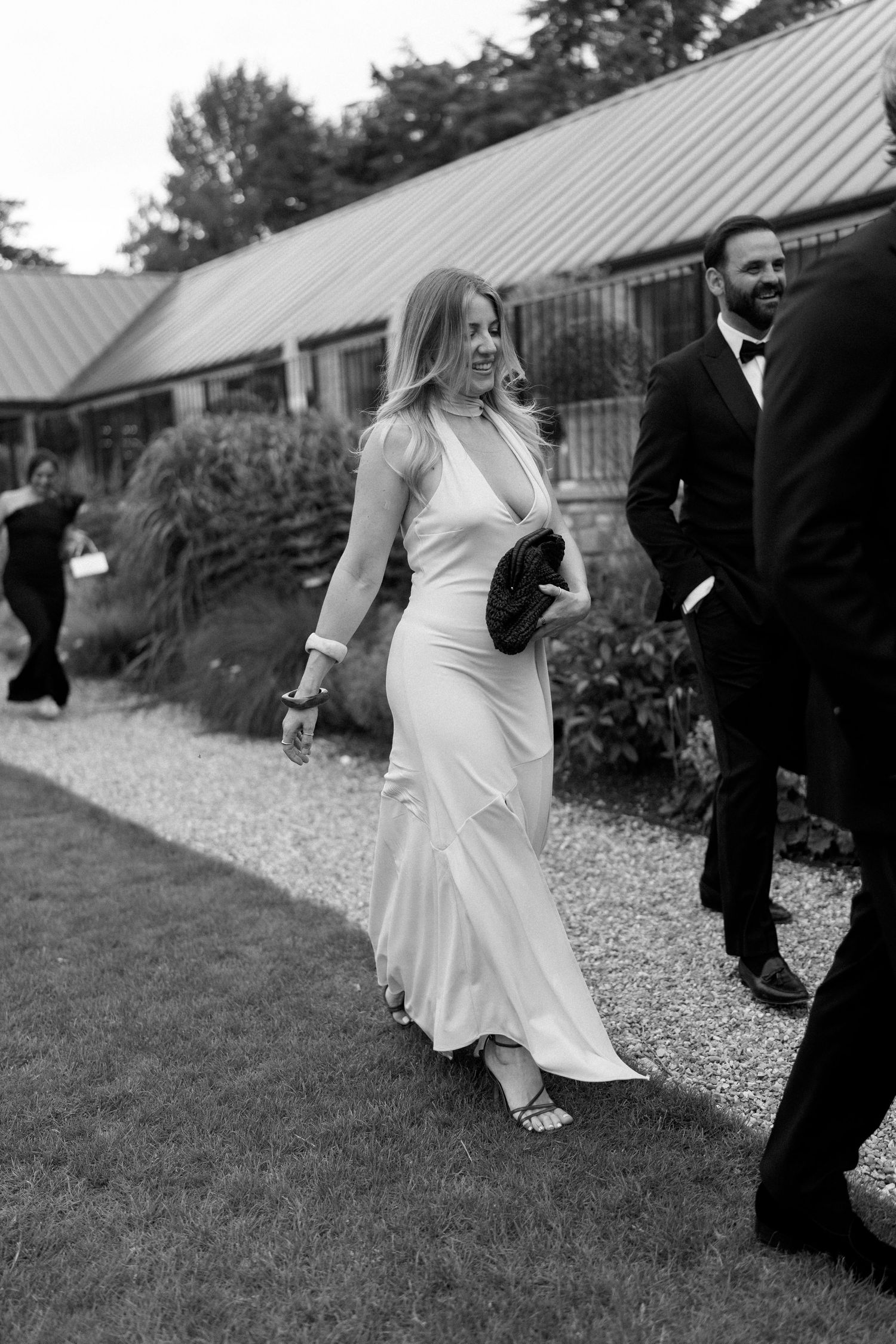 Black and white photo of bride in sleek white gown with black clutch walking with guests at outdoor wedding venue.