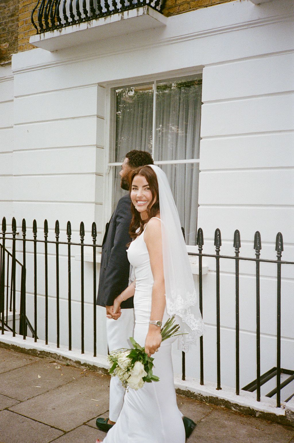 Smiling bride in white gown and veil holds bouquet while groom in dark suit walks beside iron fence.