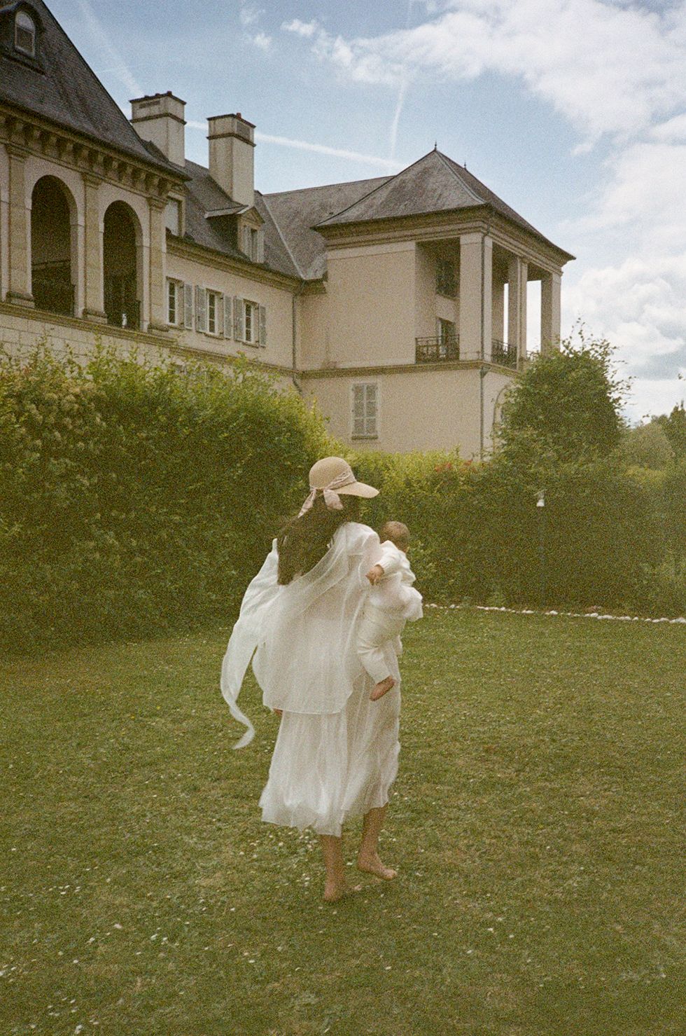 A person in a white dress carries another on their back in a lush garden beside an elegant French manor.