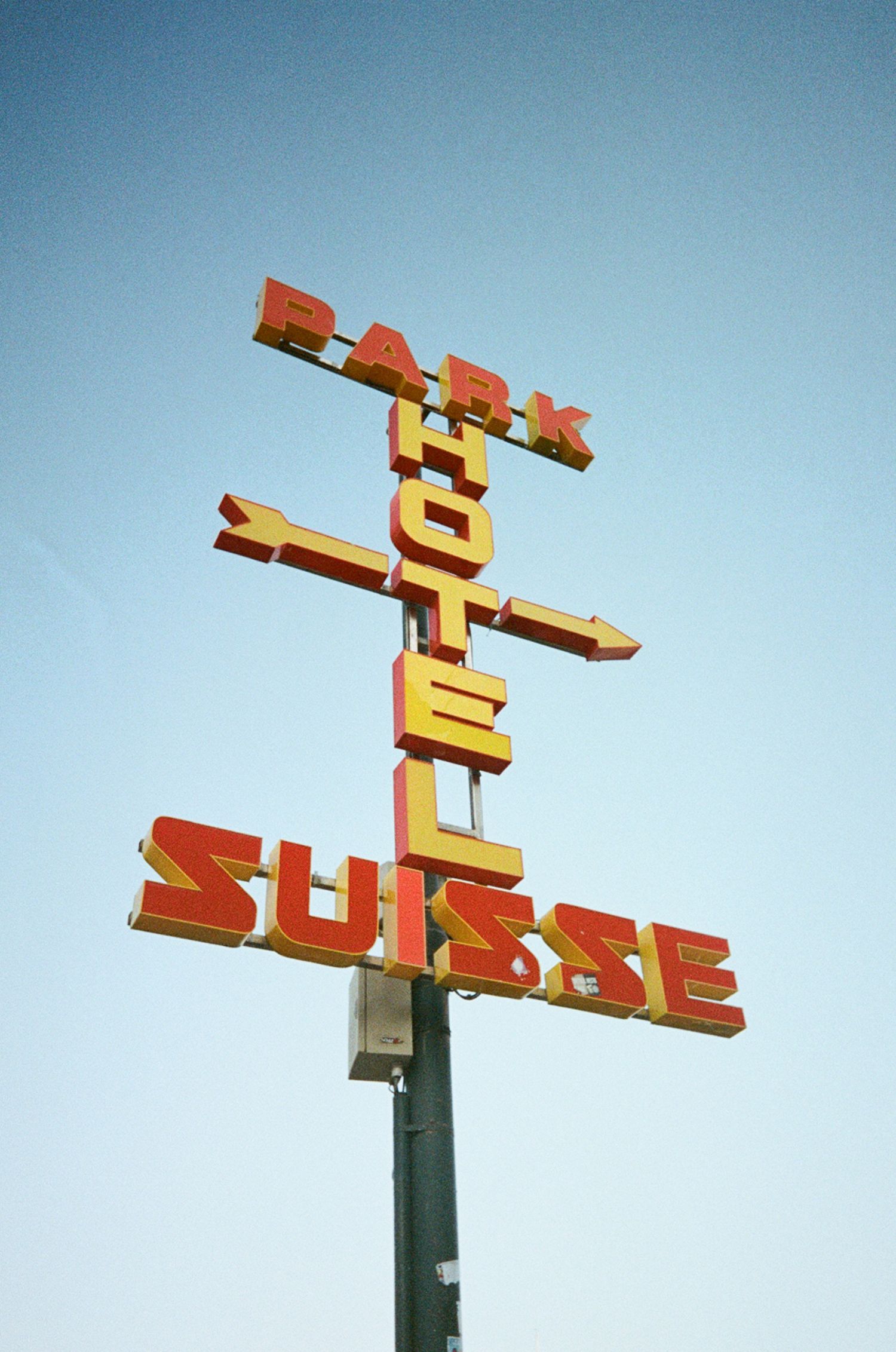 Vintage red and yellow neon sign for Park Hotel Suisse mounted on a pole against a clear blue sky.