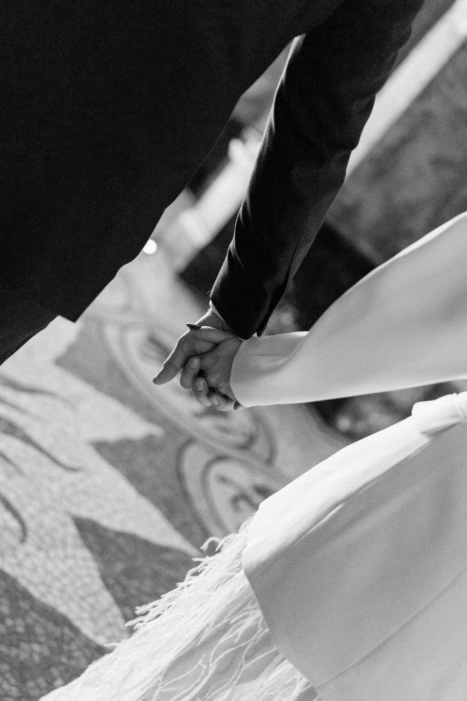Black and white close-up of bride and groom holding hands during wedding ceremony on ornate patterned floor.