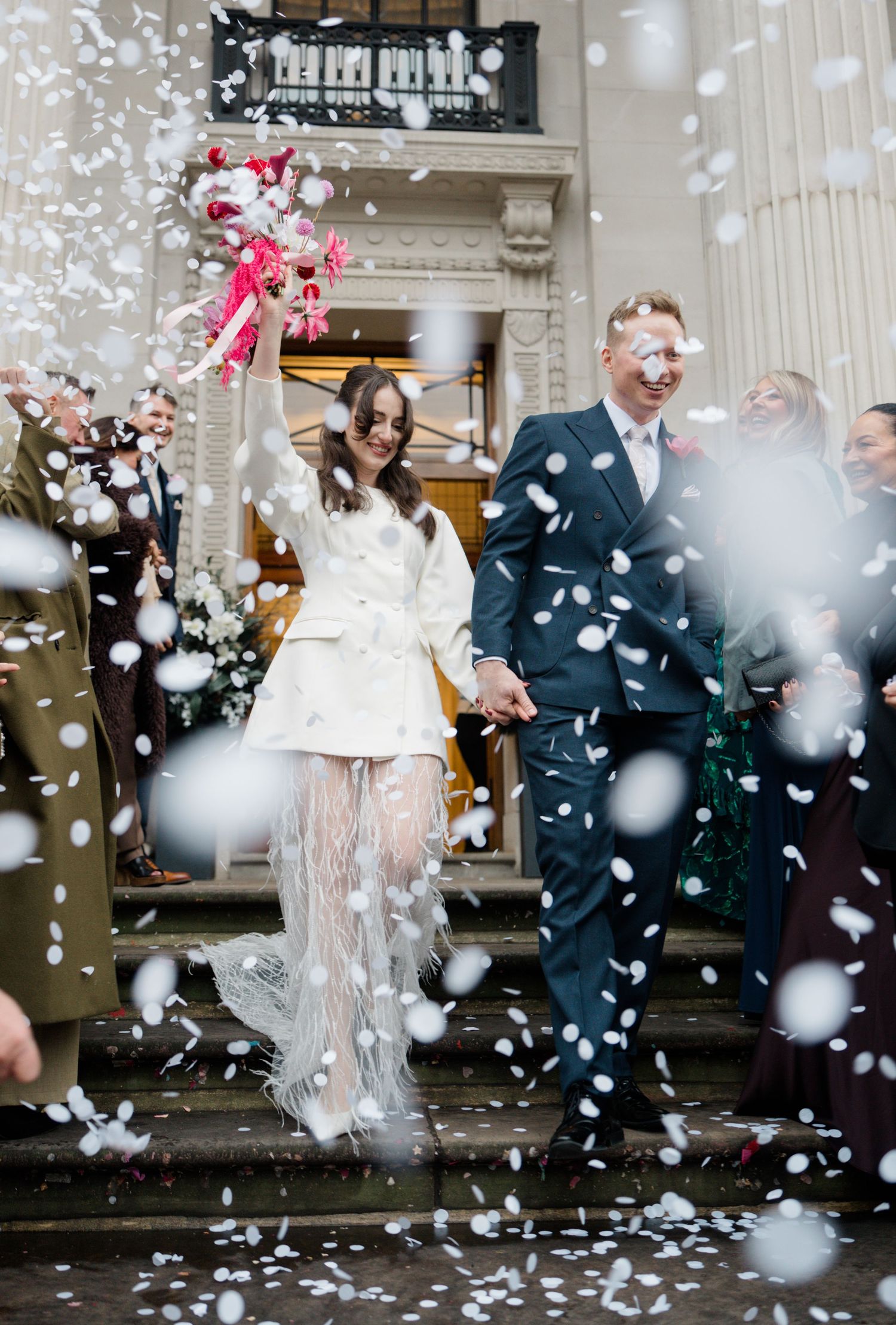 Bride and groom exit building in confetti shower, bride holds bouquet aloft.