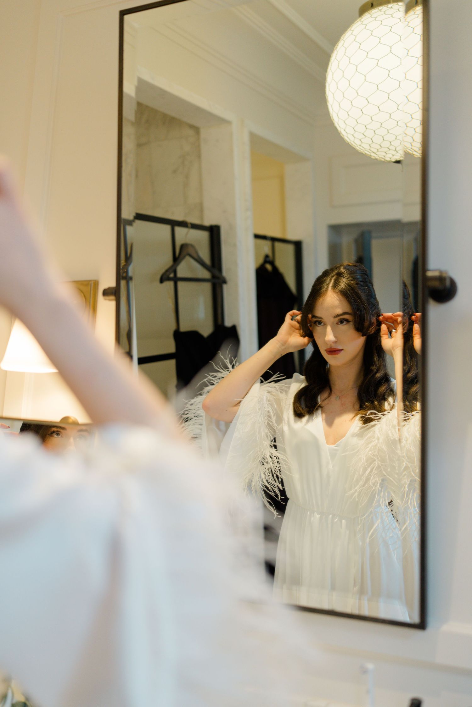 Brunette bride in white feather robe adjusting earring while gazing into elegant mirror.