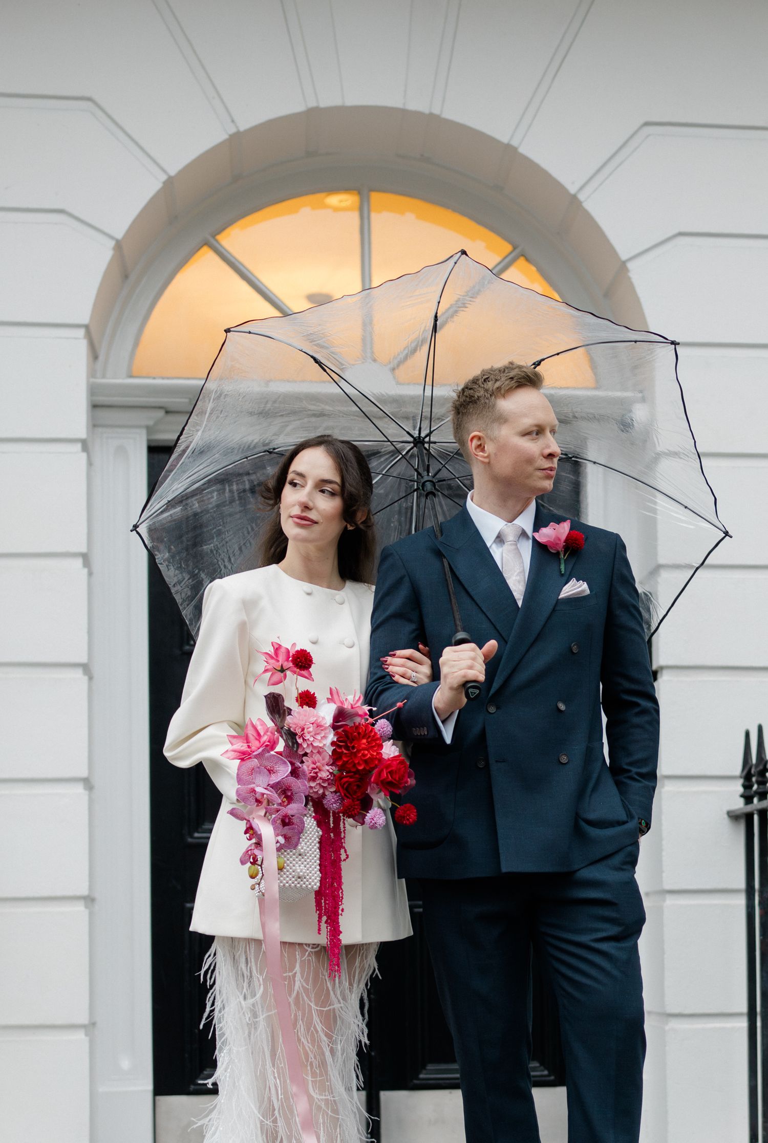 Bride and groom sharing clear umbrella outside white Georgian building with pink and red bouquet.