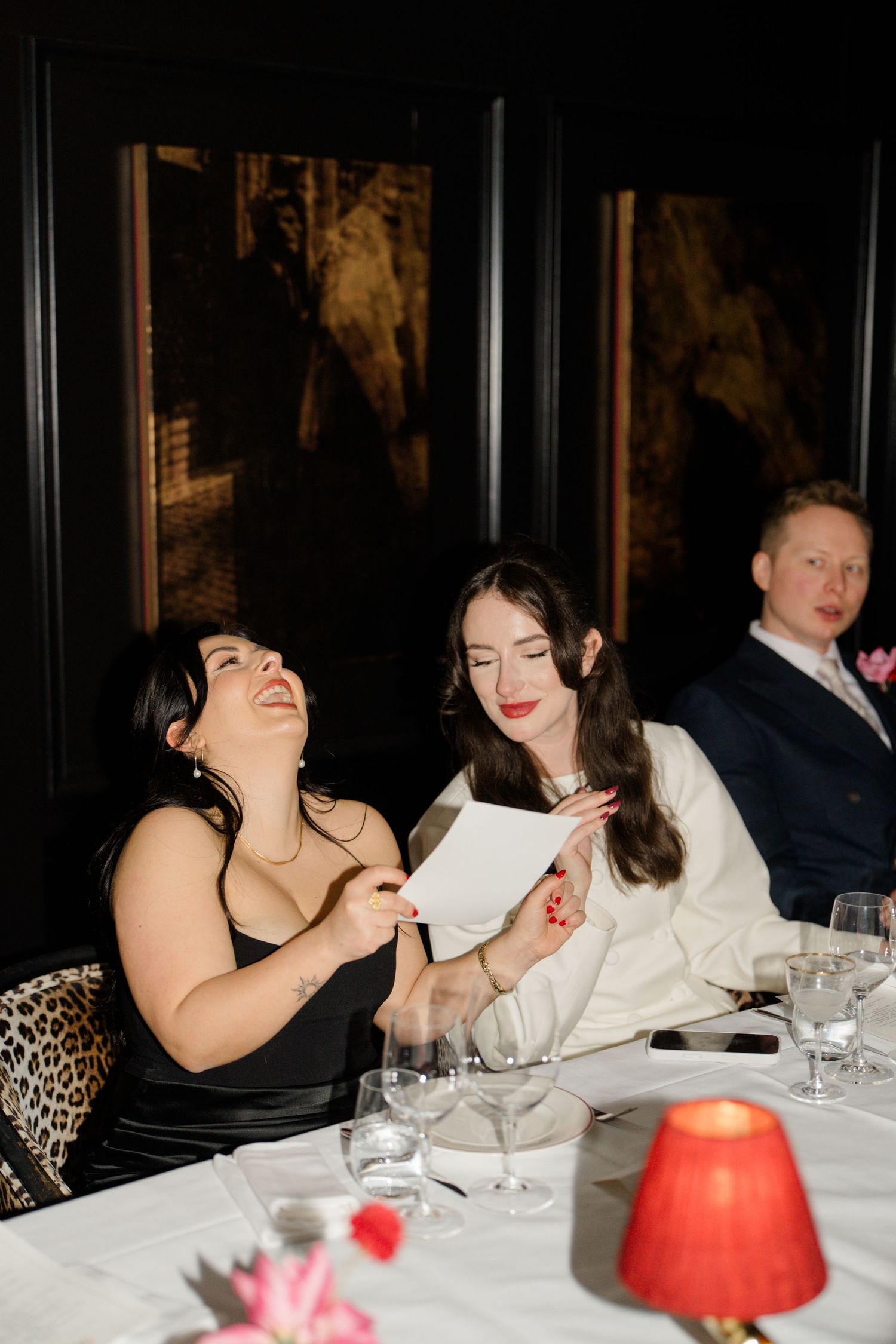 Two women laughing and reading note at wedding reception dinner table with candles.