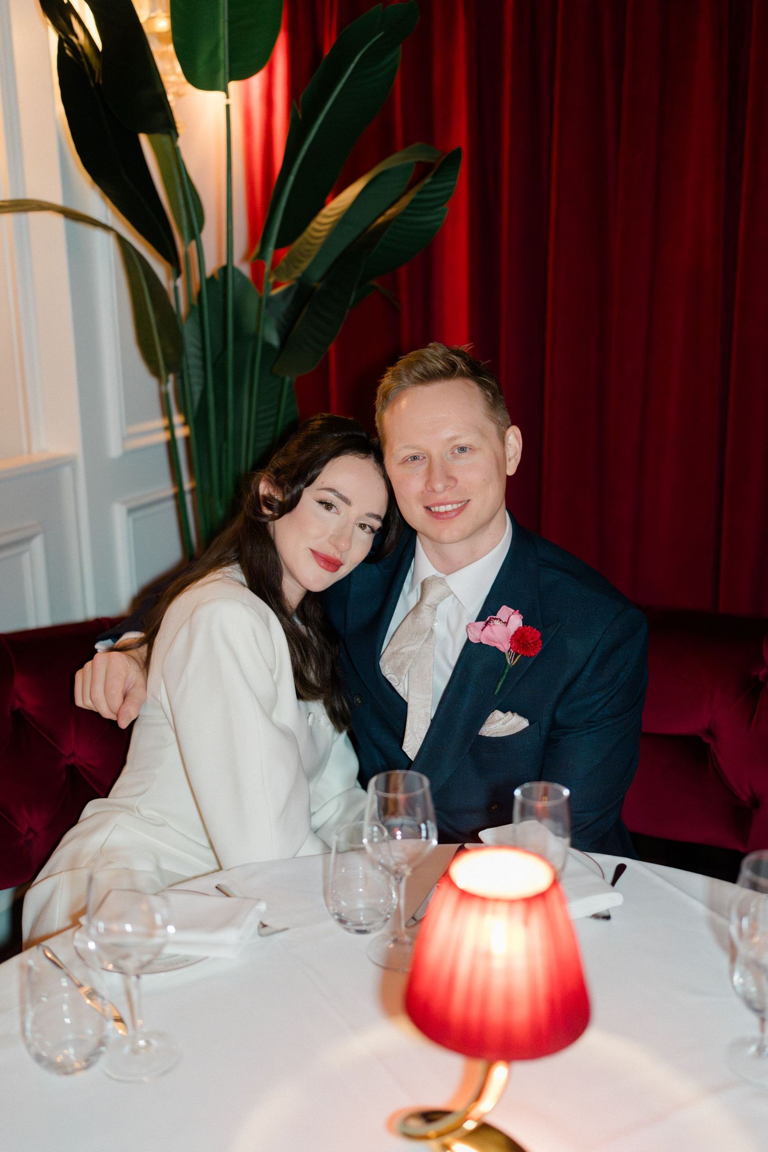 Smiling newlywed couple seated together at elegant restaurant dinner table with red lamp and velvet drapes.